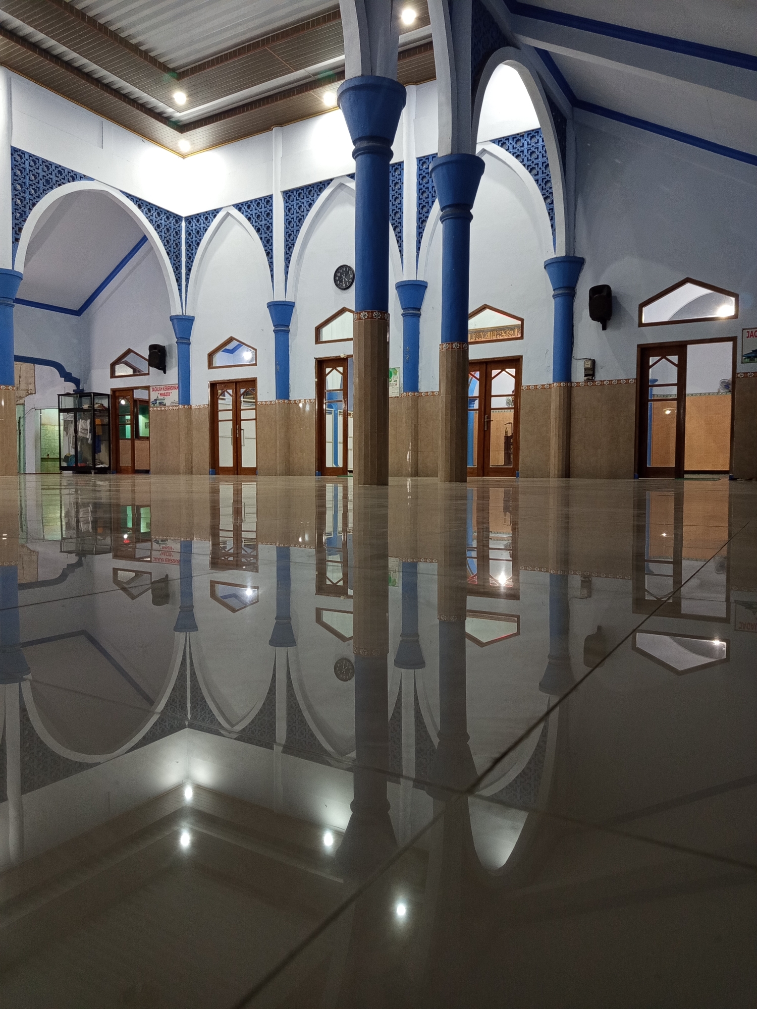 The image shows the interior of a mosque in Ngablak, Indonesia. The scene is captured from a low angle, highlighting the highly reflective tiled floor which mirrors the architectural features of the mosque. The mosque has a light blue and white color scheme, with blue columns supporting arched ceilings decorated with intricate blue patterns. Several wooden doors are visible along the back wall, some with windows. The lighting appears to be artificial, with overhead lights casting a warm glow and creating reflections on the floor. There are no people visible in the image. The overall impression is one of quiet solemnity and architectural beauty, emphasized by the mirroring effect of the polished floor.
