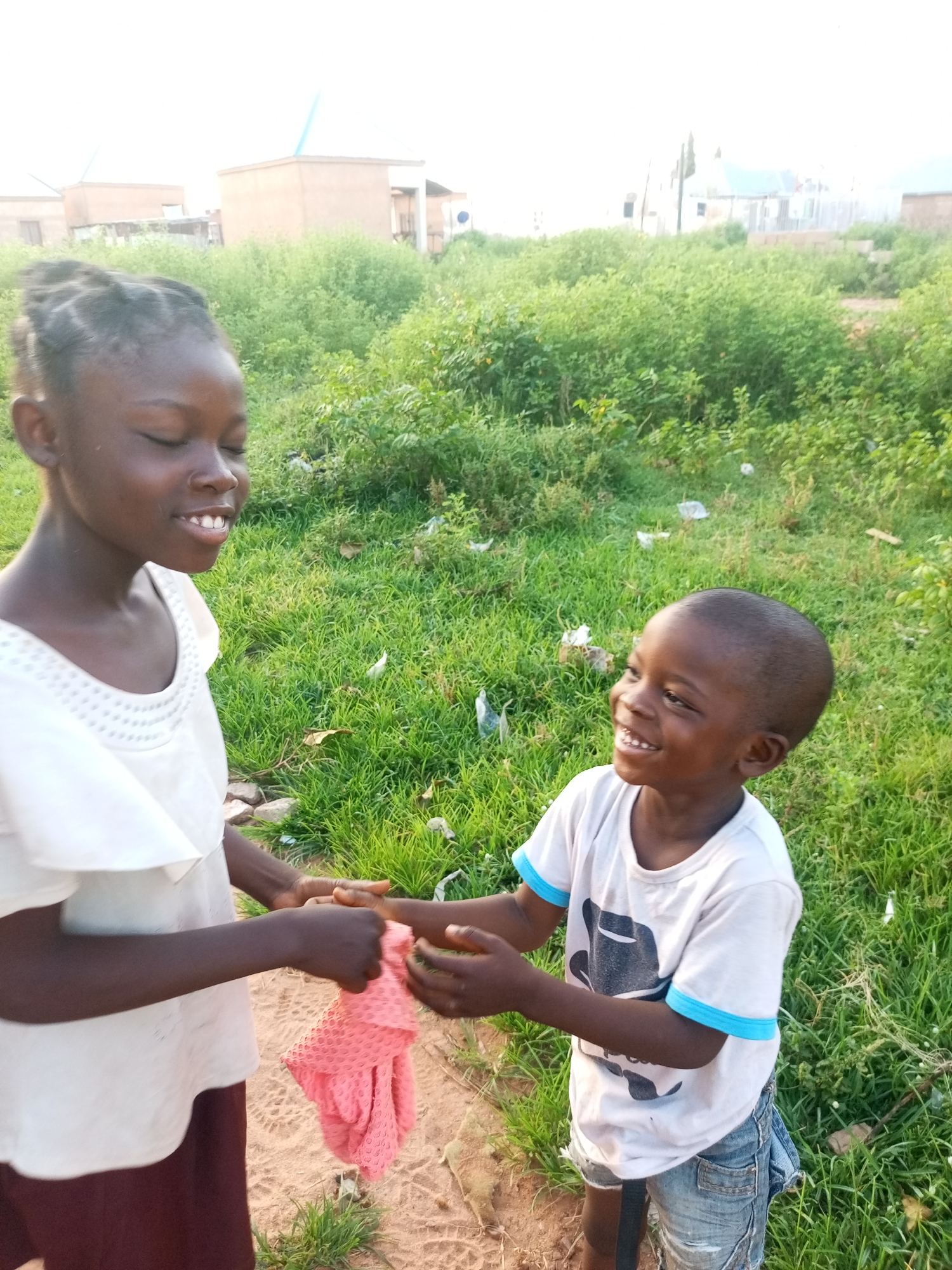 Two Black children, a girl and a younger boy, interact outdoors in a grassy, undeveloped area. The girl, on the left, has her eyes closed and is smiling. She wears a white top with a decorative neckline and a dark skirt. The boy, on the right, is looking up and to his left with a wide smile and his mouth open, showing teeth. He wears a white t-shirt with a graphic print and blue trim on the sleeves, and distressed denim shorts. The girl is handing a crumpled pink fabric item to the boy. The background consists of lush green vegetation, some scattered debris, and in the distance, low-rise buildings that suggest an urban or semi-urban setting. The lighting indicates it is daytime, possibly late afternoon, with soft light and some shadows. The overall impression is one of playfulness and youthful connection in an informal environment in Tempure, Nigeria.