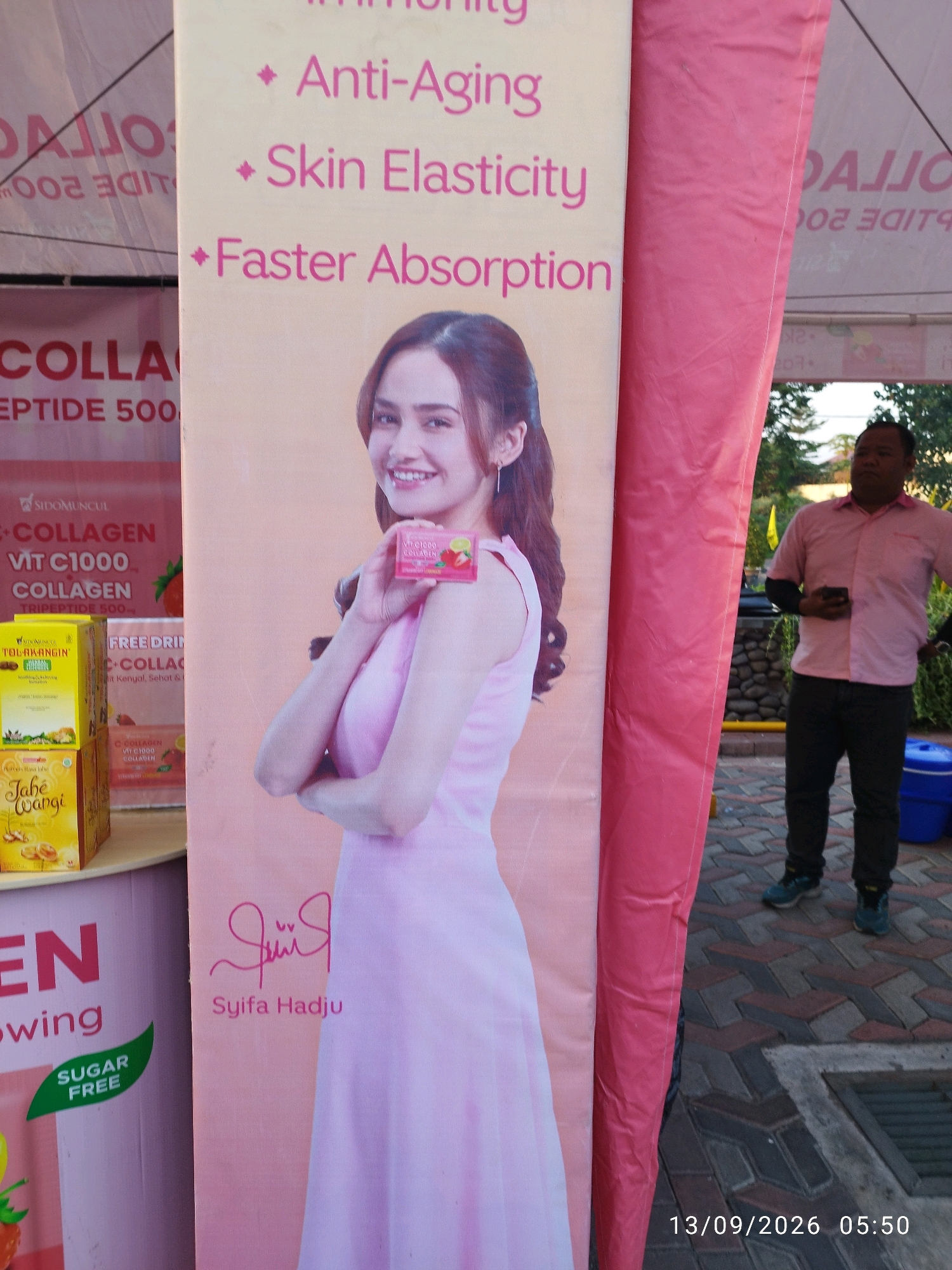 A promotional banner for collagen products is displayed vertically. The banner features a young woman in a pink dress holding a small product box, with text listing benefits like "Anti-Aging," "Skin Elasticity," and "Faster Absorption." Below her image is a signature that reads "Syifa Hadju." To the left of the banner, product boxes are visible, including "TOLAK ANGIN HERBAL ENERGI" and "Jahe Wangi." A larger banner to the far left has text that appears to be in Indonesian, possibly advertising collagen. A man wearing a pink shirt and dark pants stands to the right of the banner, holding a smartphone. In the background, there are trees and a stone wall. The scene is outdoors, with a brick-paved ground. A blue plastic bin is visible near the man. The timestamp "13/09/2026 05:50" is overlaid on the bottom right of the image. The image appears to be taken in Mojokerto, Indonesia, given the context of the displayed products.