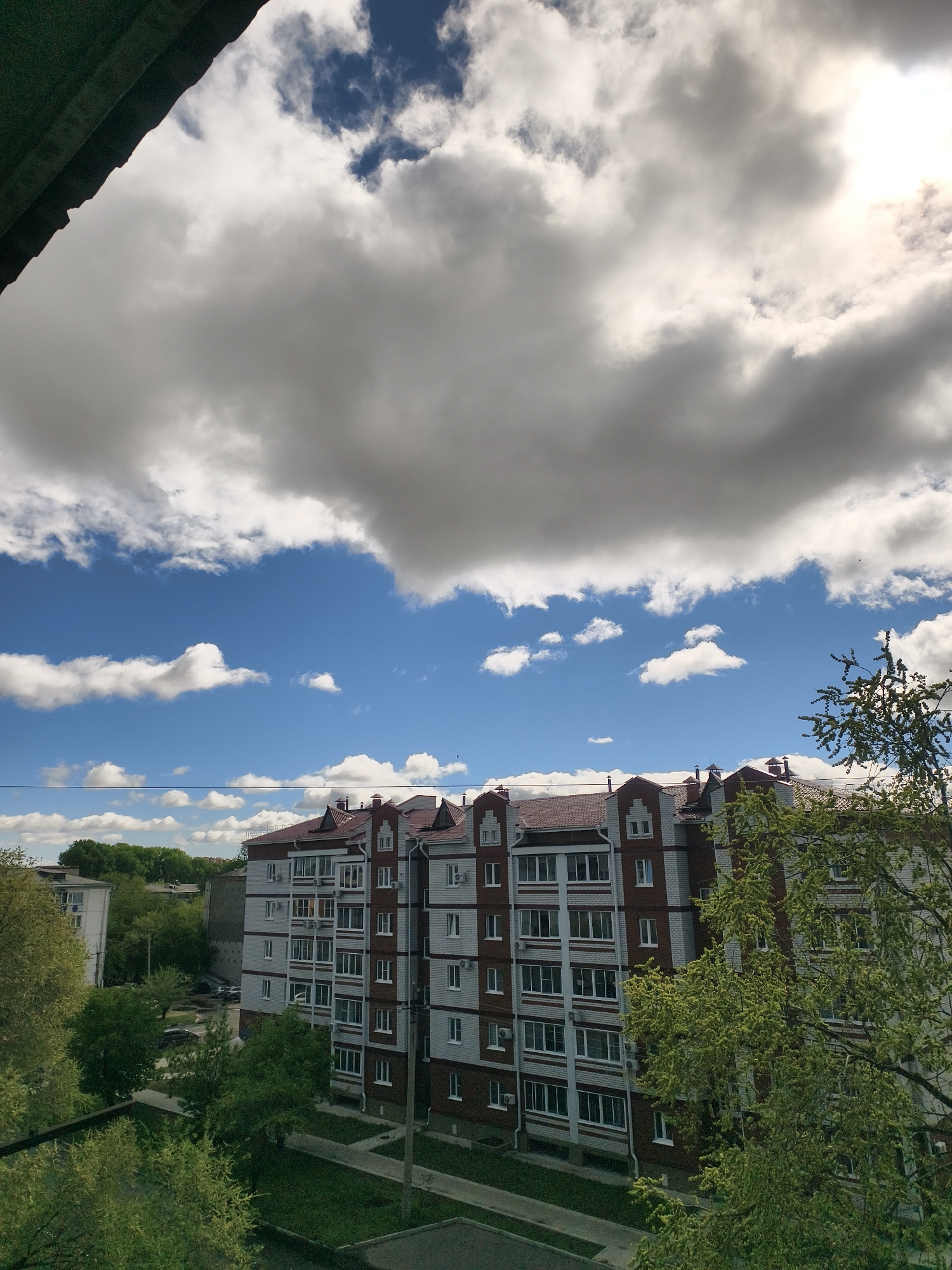 The image presents a view from an elevated position, likely a high window or balcony, in Belogorsk, Russia. The central focus is a multi-story residential building constructed of light grey brick with red brick accents and a red tile roof. White-framed windows are arranged in a regular pattern across the facade.
The building is surrounded by green foliage from trees in the foreground and along the building's periphery. A grey sidewalk is visible directly in front of the building.
The sky takes up a significant portion of the image, showcasing a mix of blue sky and large, billowing white and grey clouds. The sun is partially obscured by a cloud, casting a soft light over the scene. Part of a building is visible to the side.