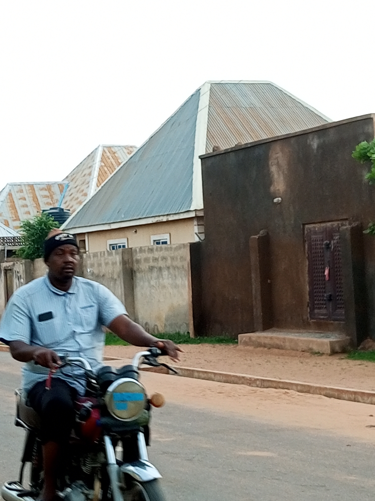A man rides a motorcycle down a dirt road in Gombe, Nigeria. The man wears a blue collared shirt and a black beanie. The motorcycle has a round headlight with blue markings and a brown accent. In the background, there are buildings with corrugated metal roofs and a dark-colored, textured wall with a wooden door. A concrete wall with square pillars runs along the side of the road, separating the road from the buildings. Green foliage is visible near the buildings. The sky is overcast and pale.