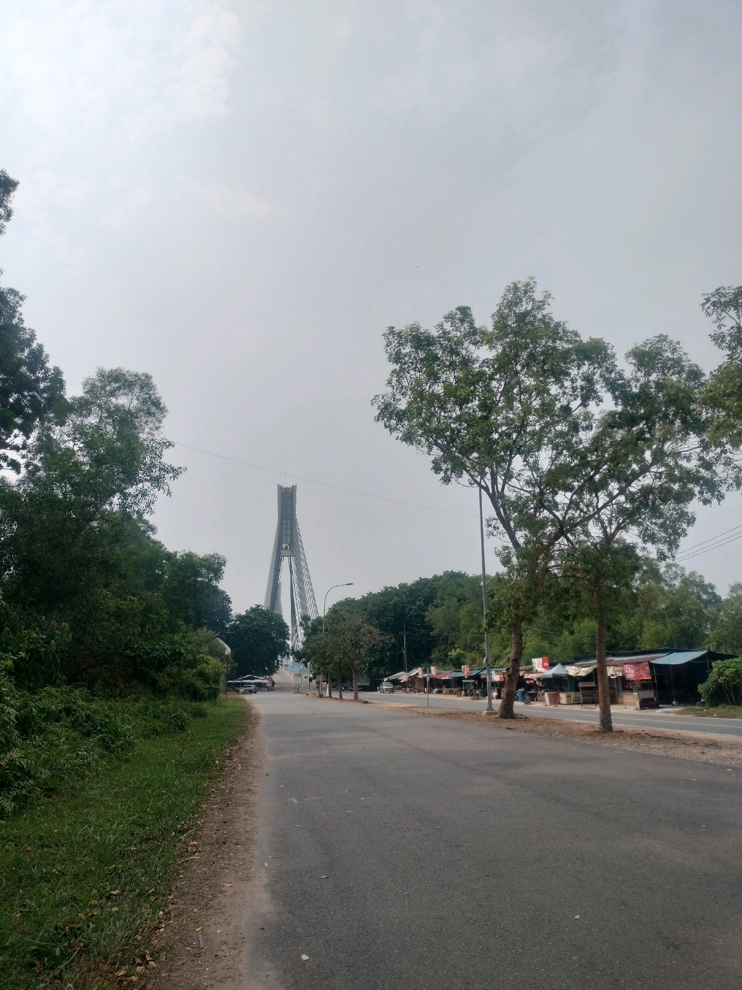 The image shows a straight, paved road with a grassy verge on the left, leading towards a large suspension bridge in the distance. The bridge has a distinctive tall, A-shaped pylon. On the right side of the road, there are several small roadside stalls or shops, some with corrugated roofs and signage. Trees line the road on both sides, with dense foliage on the left and sparser trees on the right. The sky is overcast and bright. The scene appears to be on the outskirts of a city, possibly with light traffic given the presence of a few vehicles on the road. This location is identified as the outer ring road towards Barelang Bridge in Batam, Indonesia.