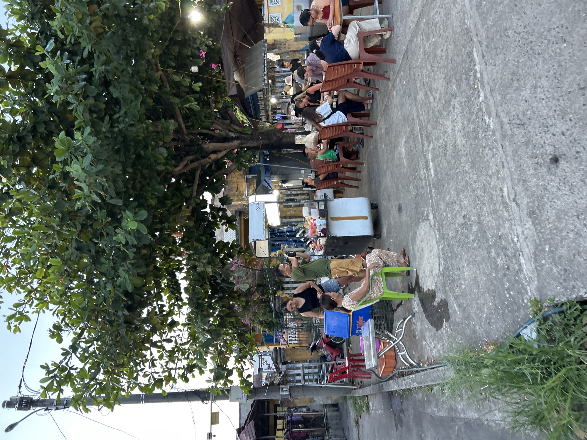 The image depicts an outdoor seating area with several people engaged in dining or conversation, likely at a food stall or restaurant in Hội An Ward, Vietnam. A large tree with dense green foliage provides shade over a portion of the scene. The setting appears to be a street or sidewalk with a concrete ground surface.
In the foreground, a woman with patterned pants sits on a green plastic chair at a metal table. To her left, a blue cooler with the "Tiger" logo is visible, next to a red chair and a small metal table with an orange bin.
Behind these elements, a group of people are seated at tables. Some individuals are actively eating or interacting with each other, while others appear to be observing their surroundings or waiting. One man, wearing a green shirt and yellow shorts, is standing near a food cart, holding a phone to his face. Another woman in a black top is standing next to him. Further back, several more individuals are seated at tables, some under a brown umbrella.
The environment suggests a casual dining atmosphere, possibly a street-side eatery or a market stall. The presence of a motorcycle on the left side of the image indicates a typical urban street scene in Vietnam. The overall lighting suggests it is either late afternoon or early evening.