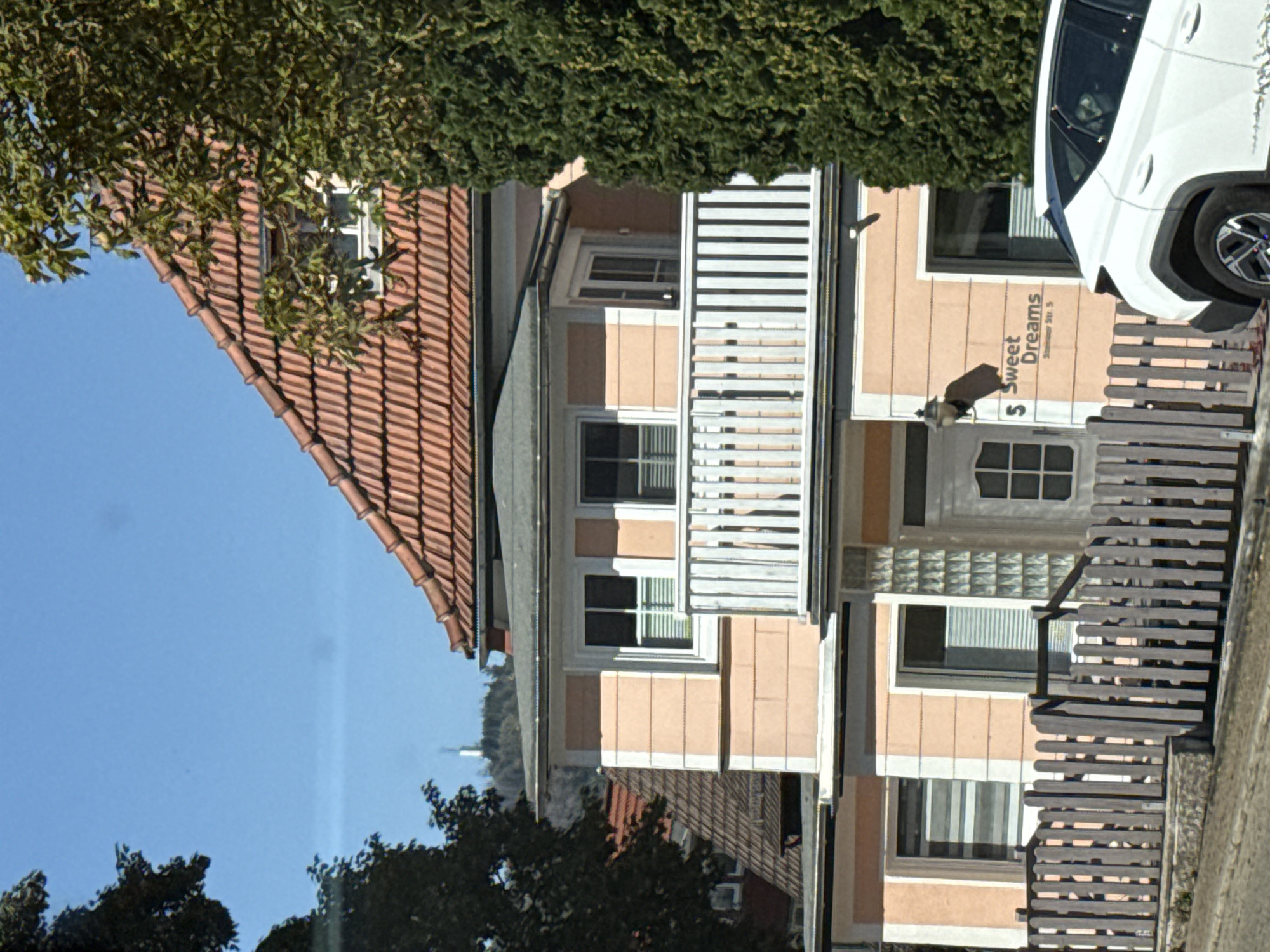 The image captures a two-story building with a salmon-pink facade and a terracotta tiled roof, located at 5 Schulstraße in Bad Sachsa, Germany. A white wooden balcony with vertical railings is visible on the second floor. The building features multiple windows, some with blinds, and a white front door with glass panes. A wooden picket fence runs along the front of the property. To the right of the entrance, a sign reads "Sweet Dreams" with the address "Schulstr. 5". A white SUV is parked in front of the fence. Lush green trees frame the right side of the building. In the background, a tall white structure, possibly a radio tower or observation tower, is visible on a distant hill. The sky is clear blue, and sunlight illuminates the scene.