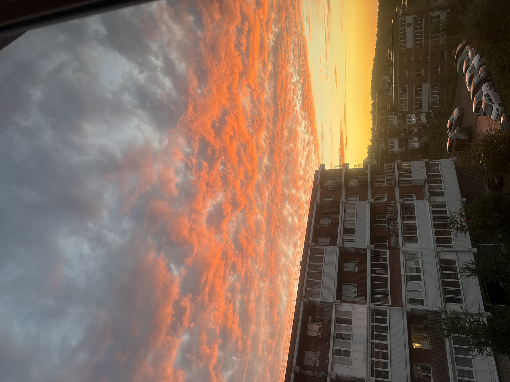 The image captures a vibrant sunset over the city of Markova, Russia. The sky is dominated by dramatic clouds painted in fiery oranges, pinks, and yellows, contrasting with darker grey clouds. In the foreground, several multi-story apartment buildings are visible. The architecture appears functional, with balconies and numerous windows. Below the buildings, a street or parking area is lined with parked cars. The overall scene depicts a residential urban environment during a spectacular natural event.