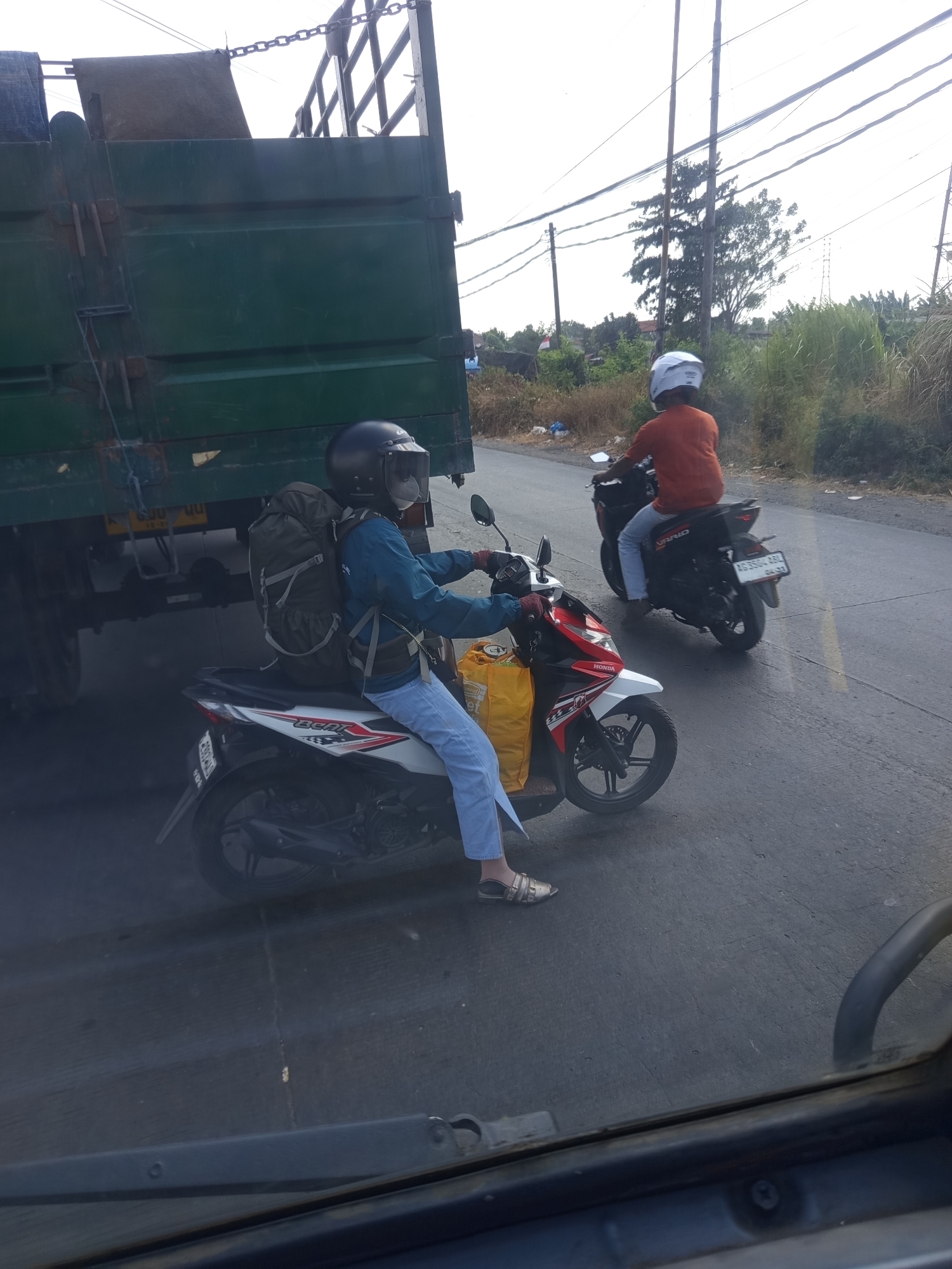 This image depicts a street scene in Sidomulyo, Indonesia, with two individuals on motorcycles. In the foreground, a person wearing a blue jacket, jeans, a helmet with a tinted visor, and a large backpack is riding a red and white scooter. The scooter has the word "BEAT" on its side. A yellow bag is strapped to the back of the scooter. In the background, another individual wearing an orange shirt and a white helmet is riding a black motorcycle. This motorcycle has a license plate that reads "AG 3584 ABL". Both individuals appear to be traveling on an asphalt road, with a large green truck to the left of the frame. The environment suggests a semi-urban or rural setting, with vegetation visible along the roadside and power lines overhead. The lighting indicates it is daytime, with some glare on the windshield in the foreground, suggesting the photo was taken from inside a vehicle.