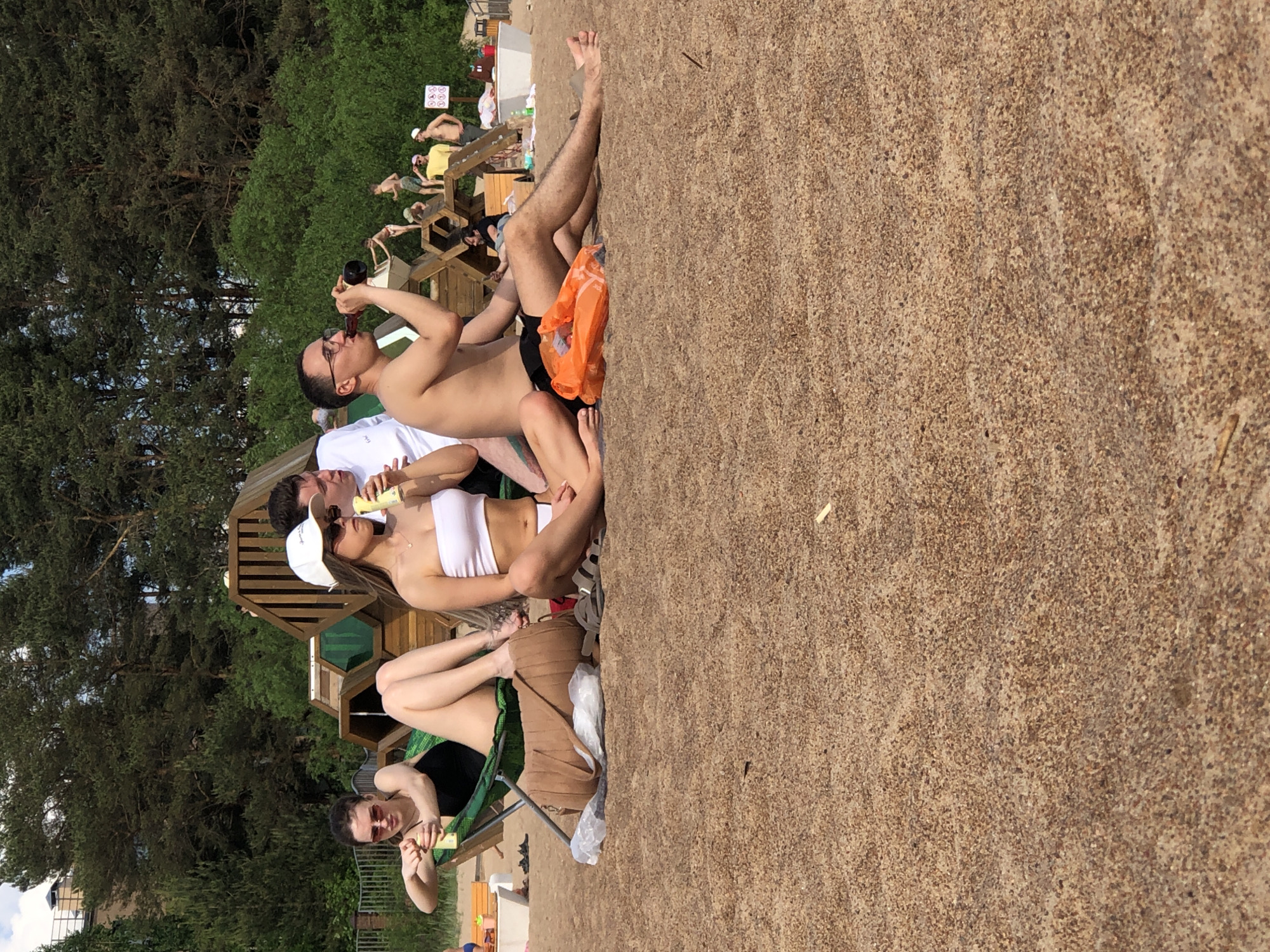 The image presents a sunlit beach scene in Sestroretsk, Russia, featuring four young adults relaxing in the foreground. On the far left, a woman with dark hair and sunglasses is seated in a green folding chair, wearing a black top, holding a yellow beverage can. Her legs are extended forward. Adjacent to her on the sand, a woman with long light brown hair, wearing a white bandeau top, white bikini bottoms, a white baseball cap, and sunglasses, holds a similar yellow beverage can. A man in a white t-shirt is partially visible behind the second woman. To the right, a shirtless man with eyeglasses and black swim shorts is seated on the sand, consuming a beverage from a dark glass bottle. His legs are extended.
In the immediate foreground, a brown suede-like handbag and a clear plastic bag containing white items are positioned on the sand near the first woman. A pair of dark sandals are visible near the second woman's feet, and an orange plastic bag containing orange items rests near the shirtless man's legs.
The mid-ground features a multi-tiered wooden structure with hexagonal and rectangular elements, some incorporating green panels, which functions as a playground or lounging area. Several other individuals are visible on and around this structure. The background consists of a dense forest of dark green coniferous trees, with a building featuring a brown roof and a light-colored facade partially visible through the foliage on the far left. A white sign with red symbols is discernible on the far right. The ground is covered in light brown, textured sand. The sky above is clear and blue.