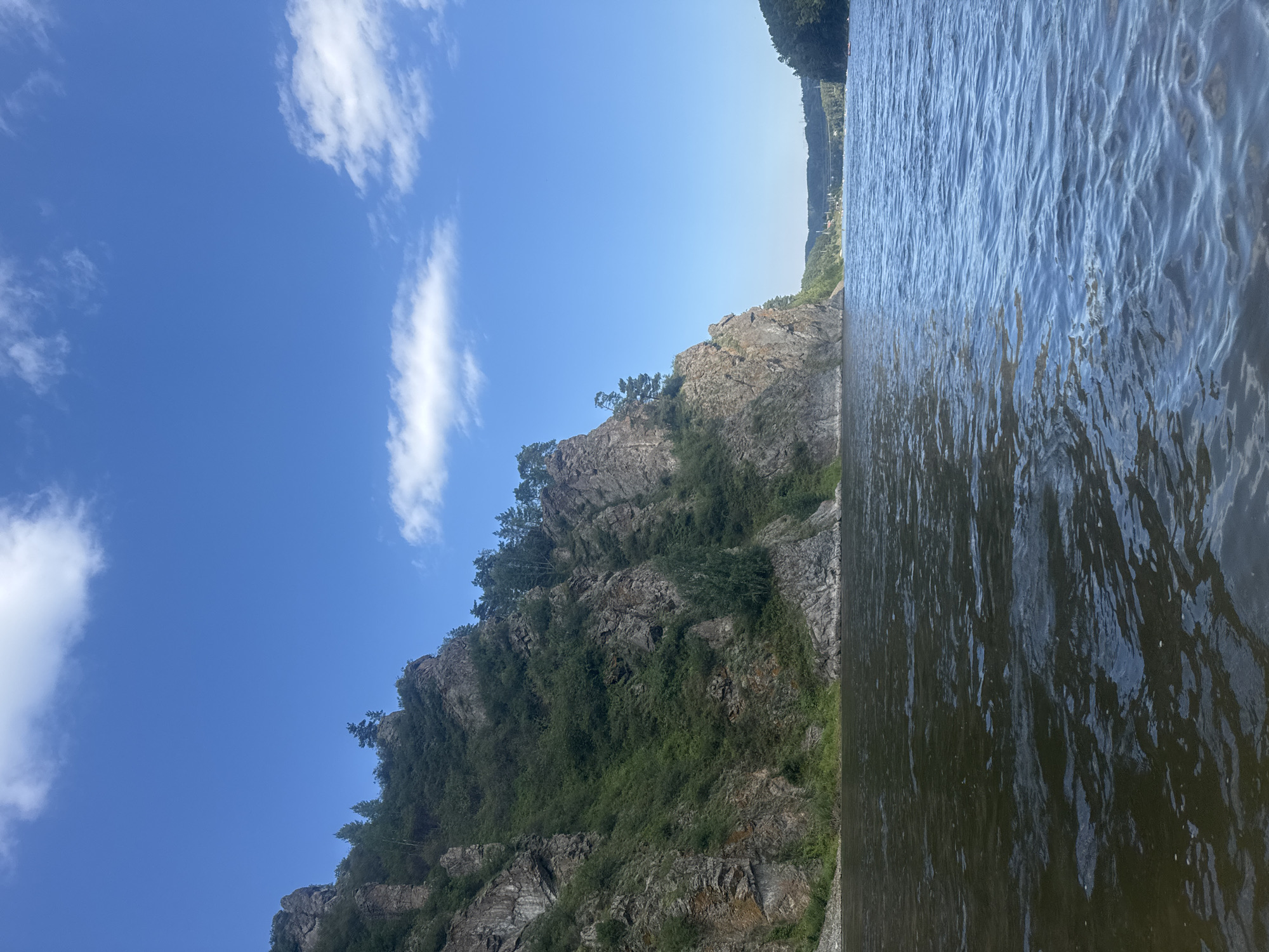 An eye-level, wide-angle shot captures a wide river with a rocky, tree-covered cliff face on the left and a forested valley in the distance on the right. The sky is a clear blue with scattered white clouds. The river is dark and reflects the surrounding environment, with ripples on the surface. The cliff face is steep and rugged, with patches of green vegetation growing on its slopes. In the distance, across the river, is a valley with dense trees, and a bridge can be seen spanning the river. The scene is likely a natural landscape in the vicinity of Ust-Mana, Russia, given the characteristic geography of the region, known for its rivers and rugged terrain. No people or specific man-made structures are clearly identifiable in the immediate foreground or on the cliff face, though distant structures suggest human presence further downriver.