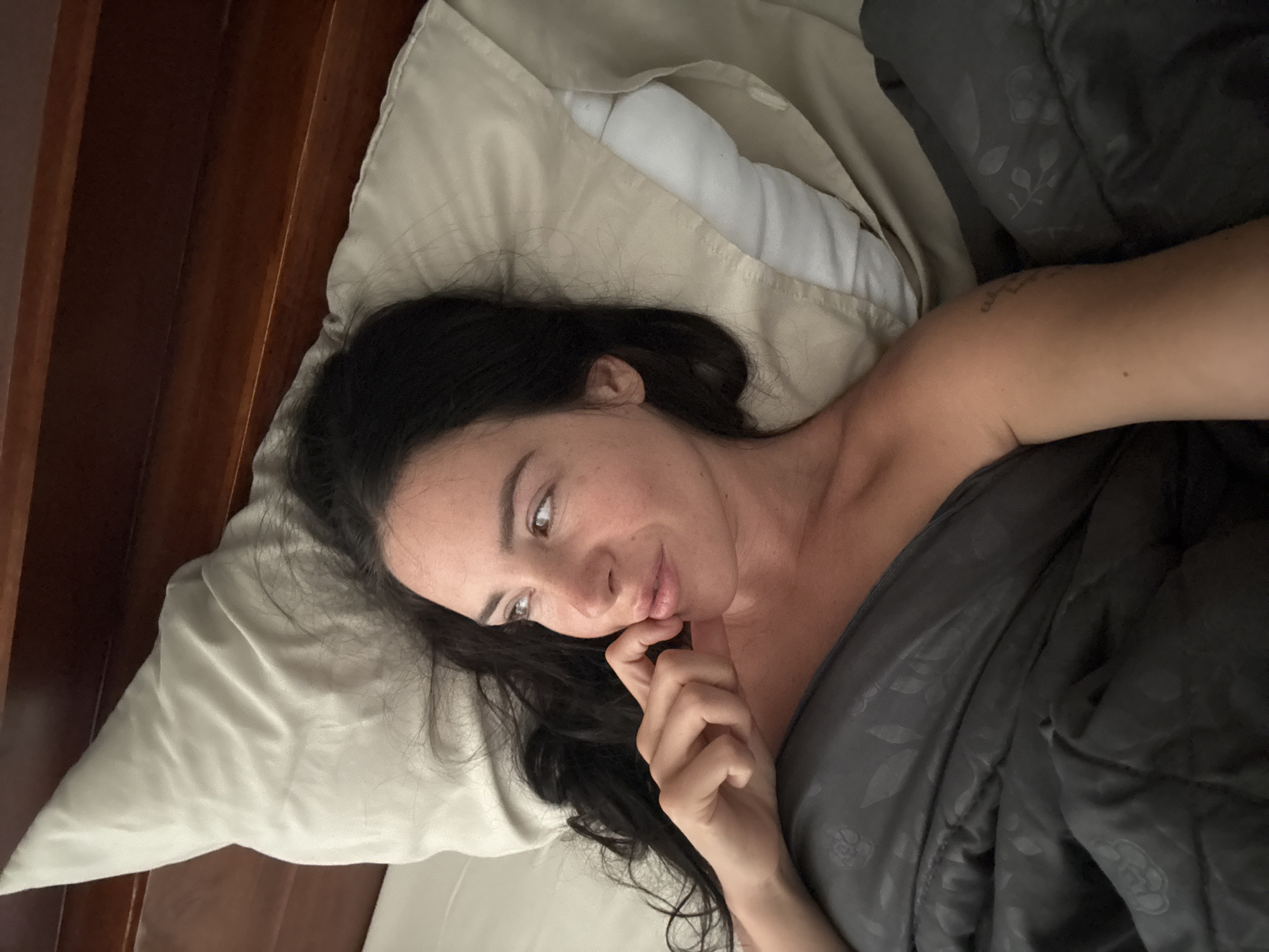 An overhead shot shows a woman lying on her side in bed, facing left and looking into the distance. Her dark hair is spread across a beige pillow, and she is partially covered by a dark grey comforter. The bed has a wooden headboard. The background suggests an indoor setting, likely a bedroom in North Kuta, Indonesia, given the natural light and possibly tropical climate implied by the room's ambiance.