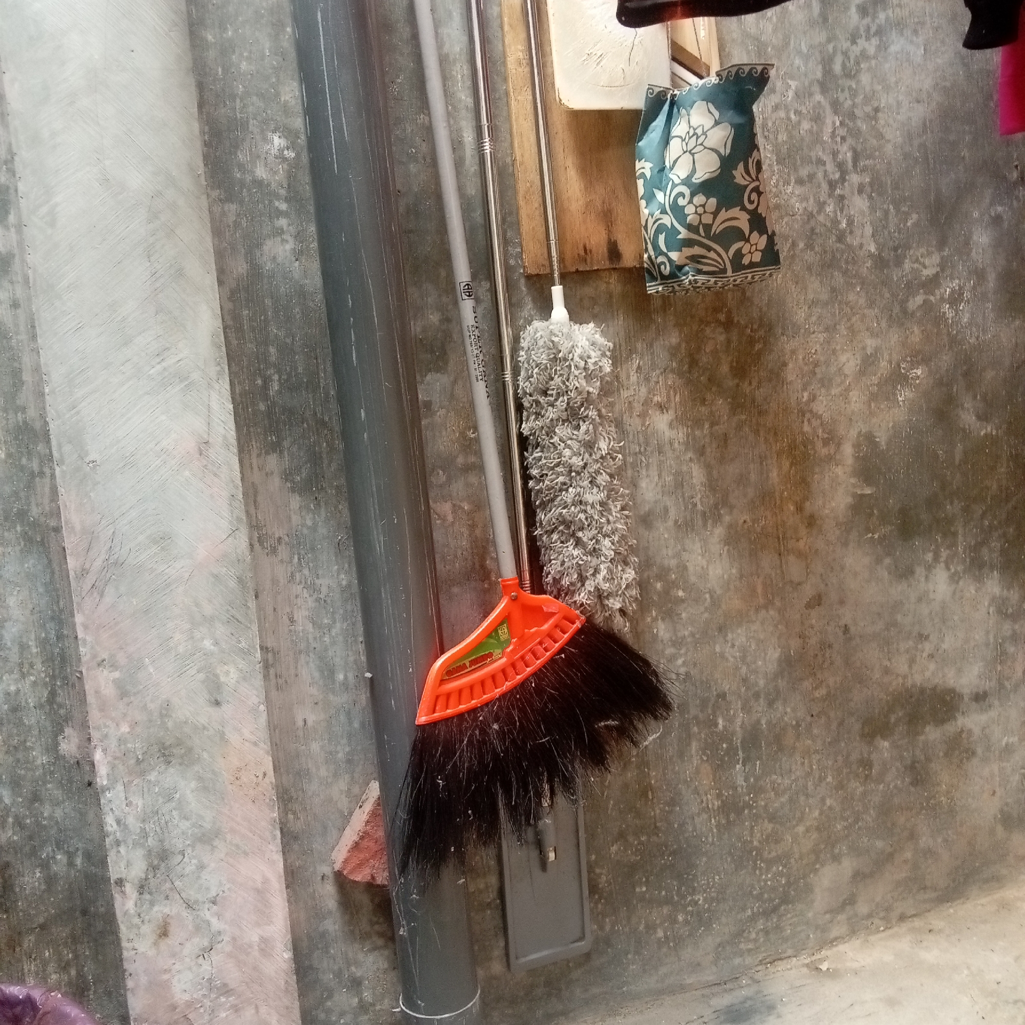 In Kediri City, Indonesia, a broom with an orange plastic head and black bristles leans against a gray pipe attached to a textured concrete wall. To the right of the broom, a gray dust mop with a fluffy head is attached to a metal pole. A decorative teal and white patterned bag hangs on the wall to the right of the dust mop. The environment appears to be an outdoor or utility area with a weathered, uneven concrete floor. A red brick piece is visible near the bottom left of the broom.