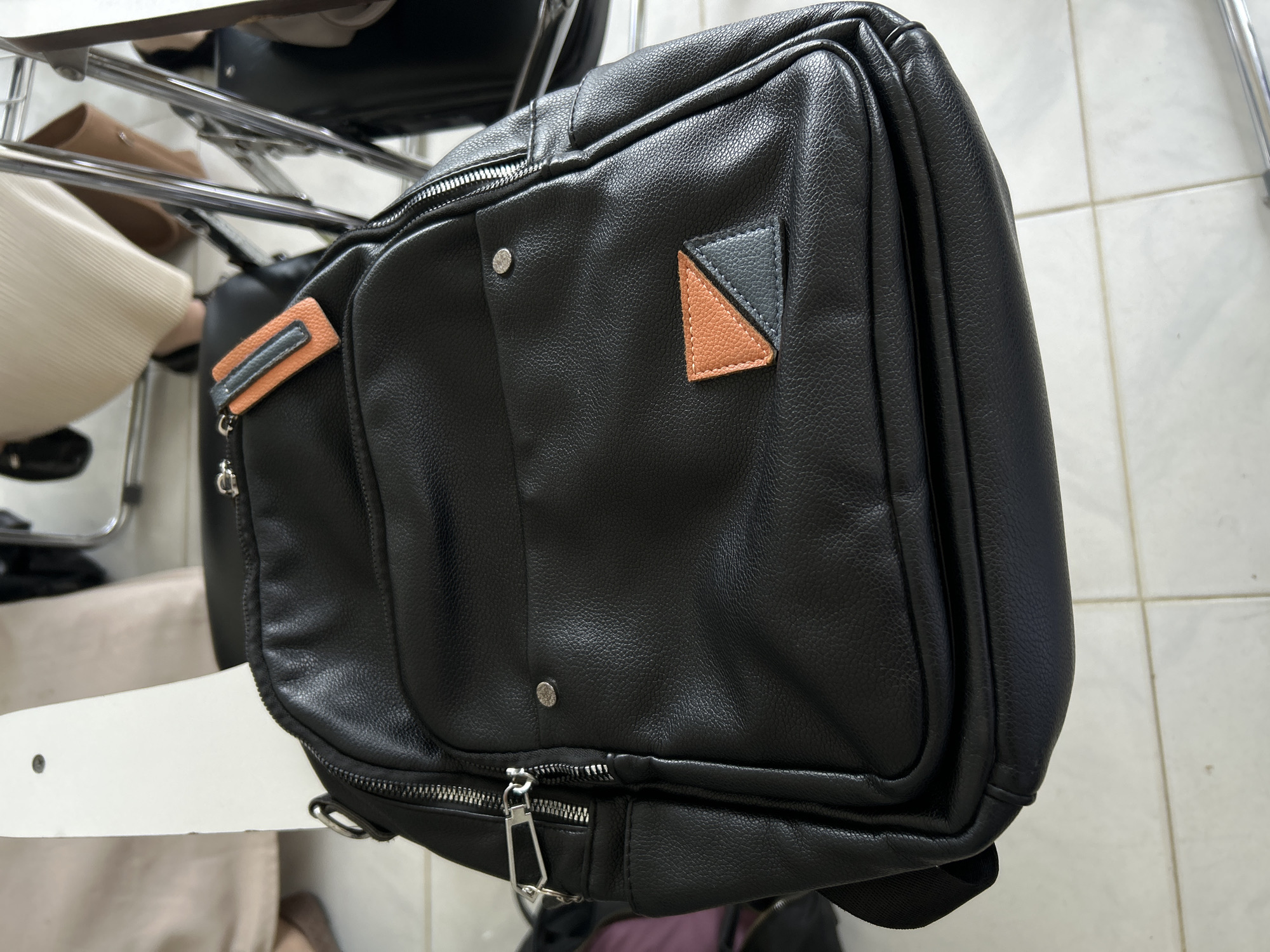 The image presents a close-up, high-angle view of a black faux leather bag, likely a messenger or shoulder bag, positioned indoors. The bag is the central subject, made of a material with a visible pebbled texture.
Notable details on the bag include multiple metallic zippers, one of which has a distinct silver-colored pull with a geometric, almost triangular base attached to a rectangular loop. Two small, round metallic studs are visible on the front panel. On the upper right side of the bag, there's a decorative patch formed by two stitched triangles: one black and one light brown or tan, outlined with light-colored stitching. To the left, a small, rectangular pull tab in light orange or tan with a greyish-blue strip is attached to a metallic ring, adorned with a small chain or charm.
The bag appears to be resting on or adjacent to a white surface, possibly a table or a chair arm. In the background, chrome-plated metal bars suggest office or household furniture. Below these bars, several blurred items are visible, including what appears to be a light brown shoe and a cream-colored ribbed fabric, possibly clothing. The floor on the right side of the frame consists of light-colored, possibly off-white or cream, square tiles with dark grout lines.
The lighting indicates an indoor setting, likely during the daytime or under bright artificial light. There is no discernible activity taking place, and no people are visible. The image focuses entirely on the static bag and its immediate surroundings. No text is visible in the frame. The scene offers no specific cues related to the provided location context of Reuleut Barat, Indonesia, beyond being an indoor environment.