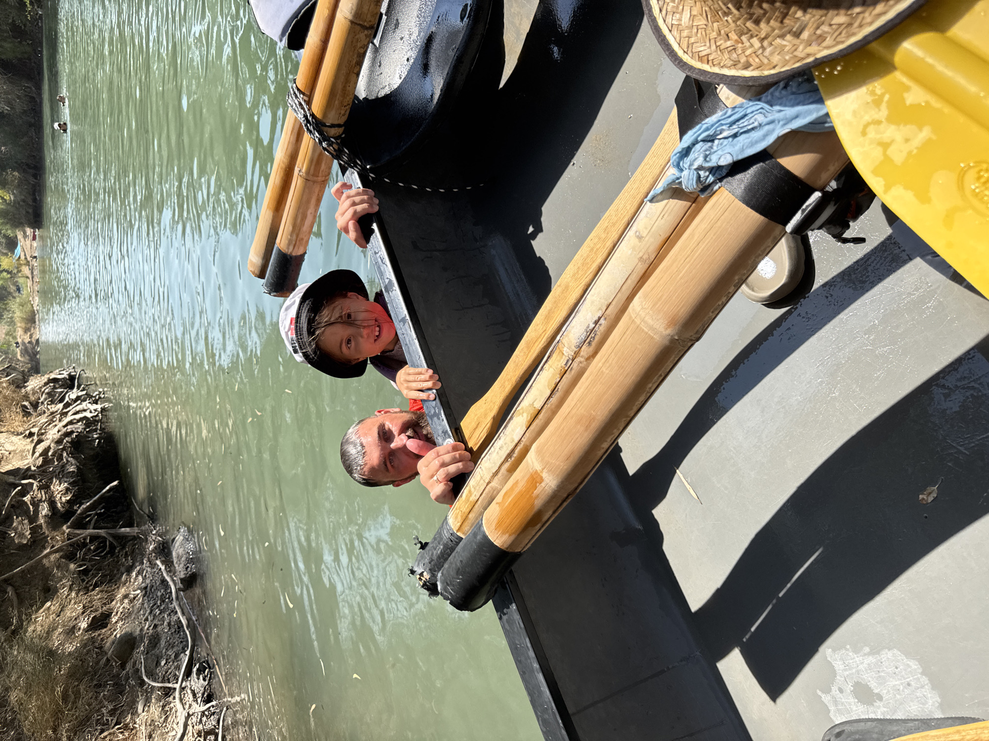 In Emek HaYarden Regional Council, Israel, on a sunny day, two individuals are partially submerged in green water while sitting in a canoe. A man with a beard and a ring on his finger is looking at the camera with a smile, with his right hand on a bamboo oar. Next to him, a child wearing a bucket hat and a life vest is also smiling at the camera, with their hands on the edge of the canoe. The canoe contains several bamboo oars secured with black straps and a blue cloth. The surrounding environment includes a muddy riverbank with visible tree roots on the left, and a lush green landscape with other people swimming in the distance on the right. The water is calm with some ripples.