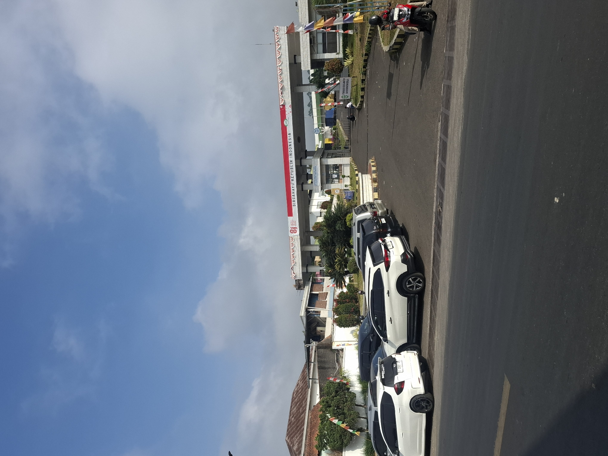 The image depicts the entrance gate of PT. Sucofindo Sukalarang in Sukalarang, Indonesia, under a partly cloudy blue sky. The gate, a substantial structure with a red and white banner that reads "Dirgahayu Republik Indonesia," is flanked by brick columns and decorated with flags. In front of the gate, several vehicles are parked, including white SUVs and a red motorcycle with a rider wearing a helmet. To the left of the gate, a building with a red tiled roof is visible, along with some trees and flags. The foreground shows a paved road with yellow lines. The scene appears to be a daytime outdoor shot, likely taken from the roadside.
