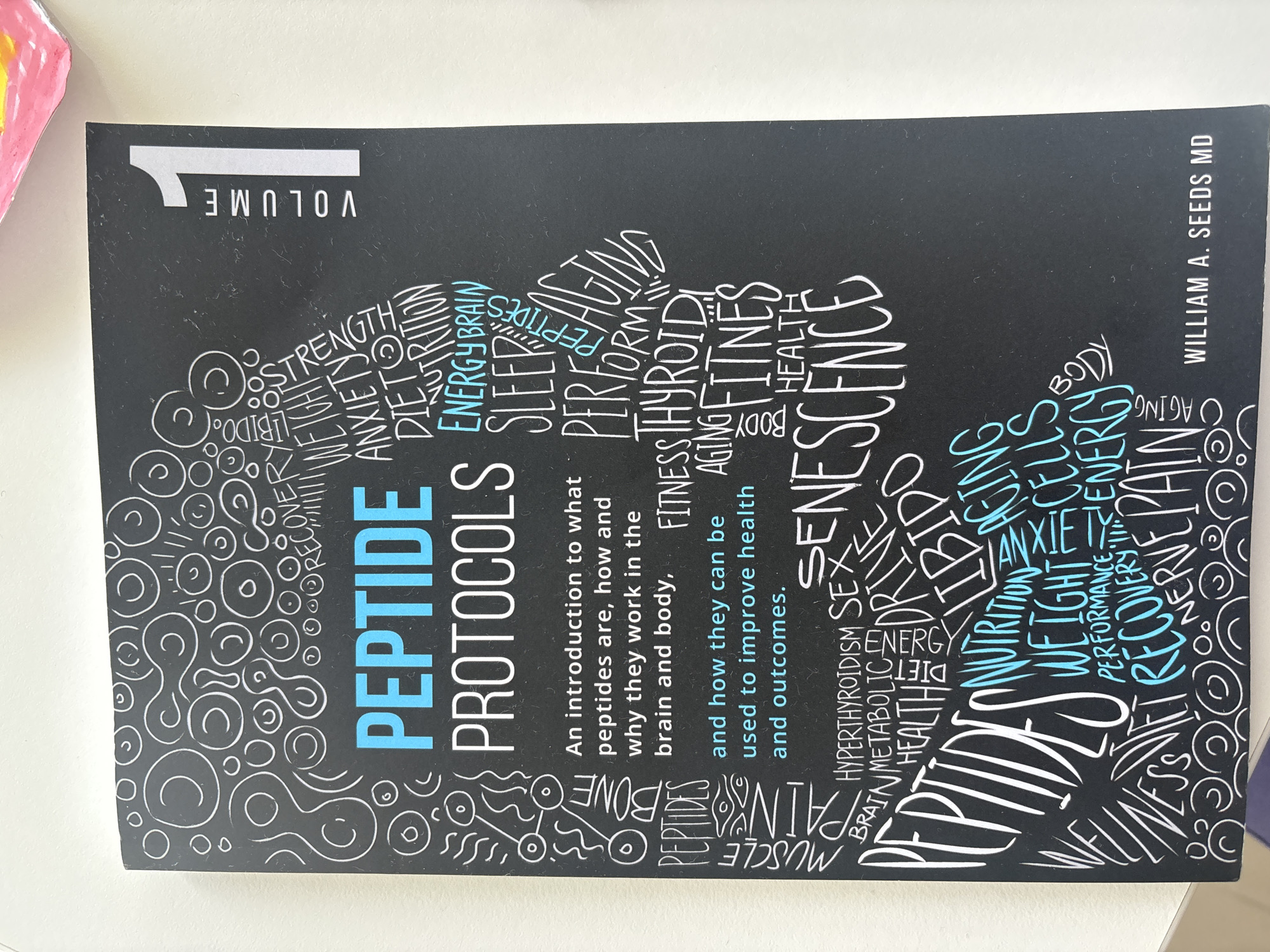 The image shows the cover of a book titled "PEPTIDE PROTOCOLS VOLUME 1" by WILLIAM A. SEEDS MD. The cover is black with white and blue text and graphics. The main title is in large blue letters, with "PROTOCOLS" in white letters below it. The graphics are a silhouette of a head and body formed by various words related to peptides, health, and aging. These words include "RECOVERY," "WEIGHT," "STRENGTH," "ANXIETY," "DIET," "NUTRITION," "ENERGY," "BRAIN," "SLEEP," "PERFORM," "AGING," "FITNESS," "THYROID," "BODY," "HEALTH," "SENESCENCE," "HYPERTHYROIDISM," "SEX," "DRIVE," "LIBIDO," "METABOLIC," "BRAIN," "MUSCLE," "PAIN," "BONE," "WELLNESS," "RECOVERY," "NERVE," and "PAIN." The word "VOLUME" appears vertically along the right side of the cover, with a large "1" next to it. The book is lying flat on a light-colored surface, possibly a table. The background is white, suggesting an indoor setting. There is no indication of the location being in Dubai, United Arab Emirates.