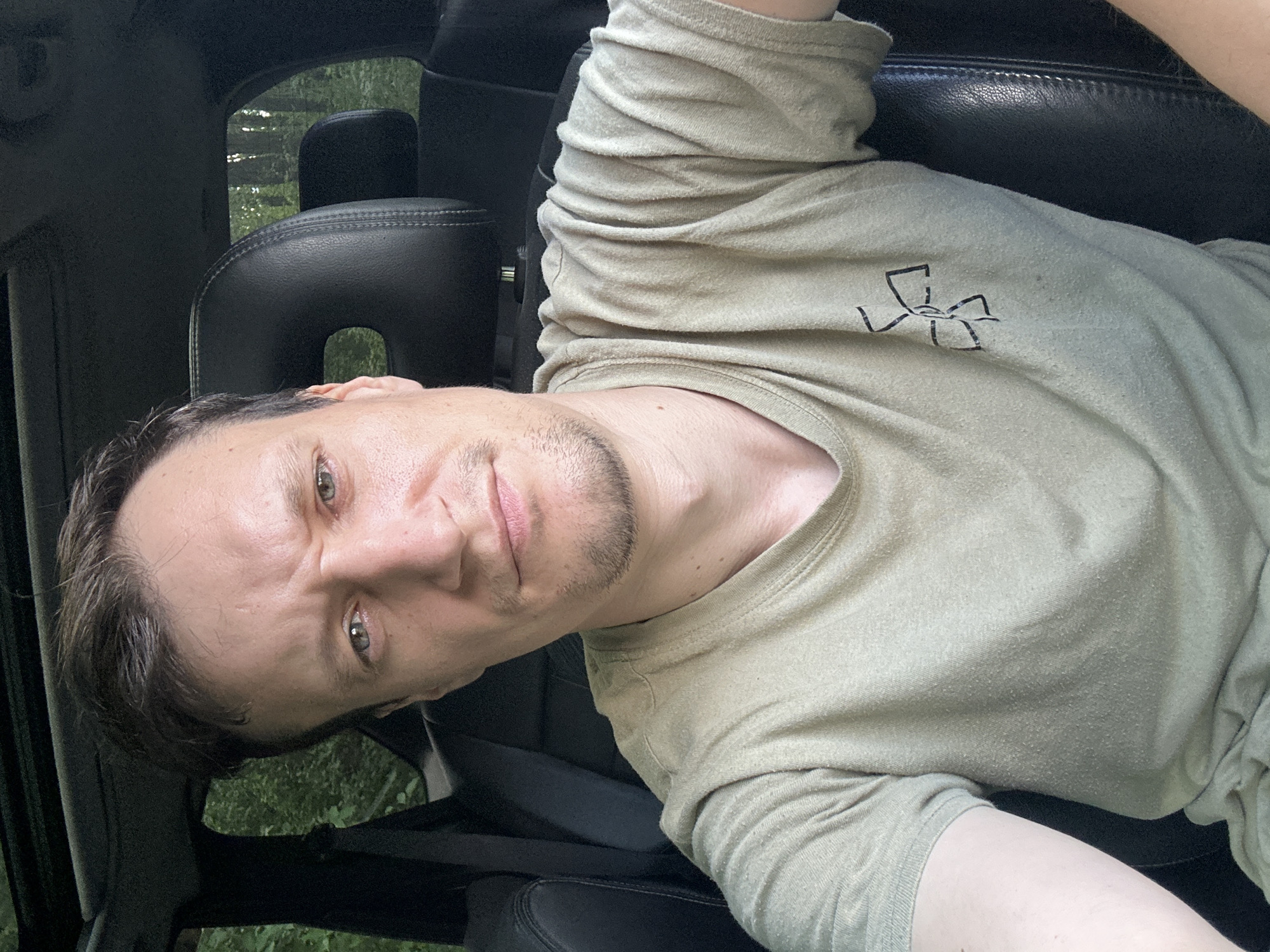 A man with light skin, dark brown hair, and blue-green eyes is sitting in the driver's seat of a car. He is wearing a light green t-shirt with a black design on the left breast. The man is looking directly at the camera with a serious expression. The car interior is visible, including the headrest and a portion of the dashboard. Outside the car, trees are visible, suggesting a natural setting. The image appears to have been taken during the daytime.