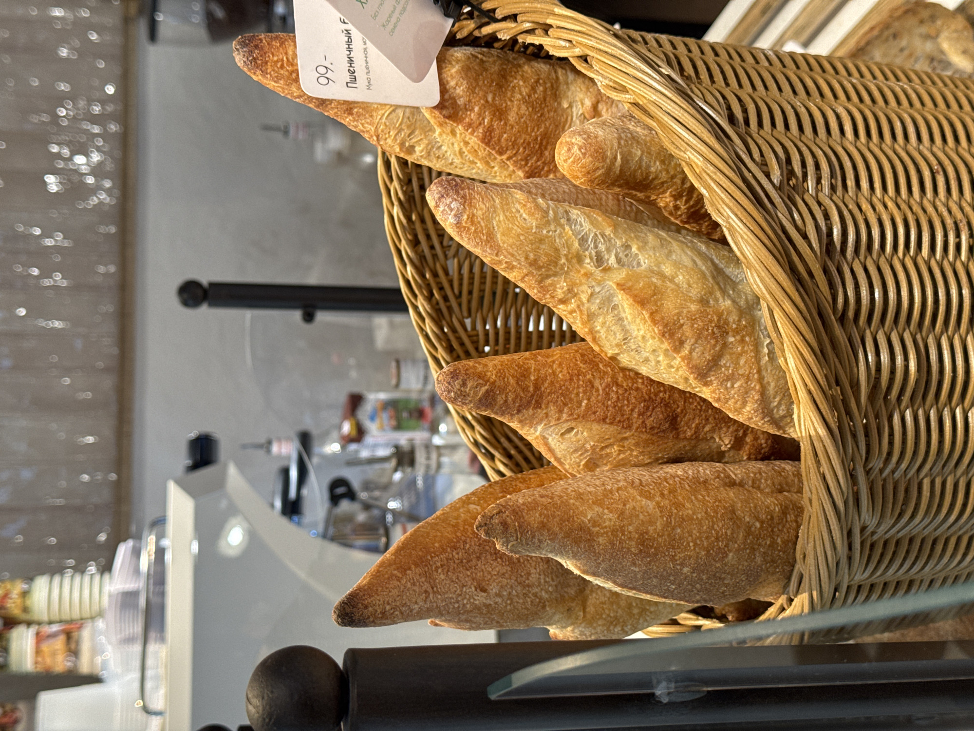 The image depicts a basket filled with baguettes in a bakery, likely located in Izhevsk, Russia, based on the Cyrillic text on a price tag attached to one of the loaves, which reads "Пшеничный F" ("Wheat F"). The basket, made of woven light brown material, is overflowing with several freshly baked baguettes, their golden-brown crusts suggesting a recent baking. The basket sits on a dark brown, likely wooden, display shelf. Background elements include a blurred view of a stainless-steel display case and other bakery items, further indicating the location is a bakery or deli within Izhevsk.