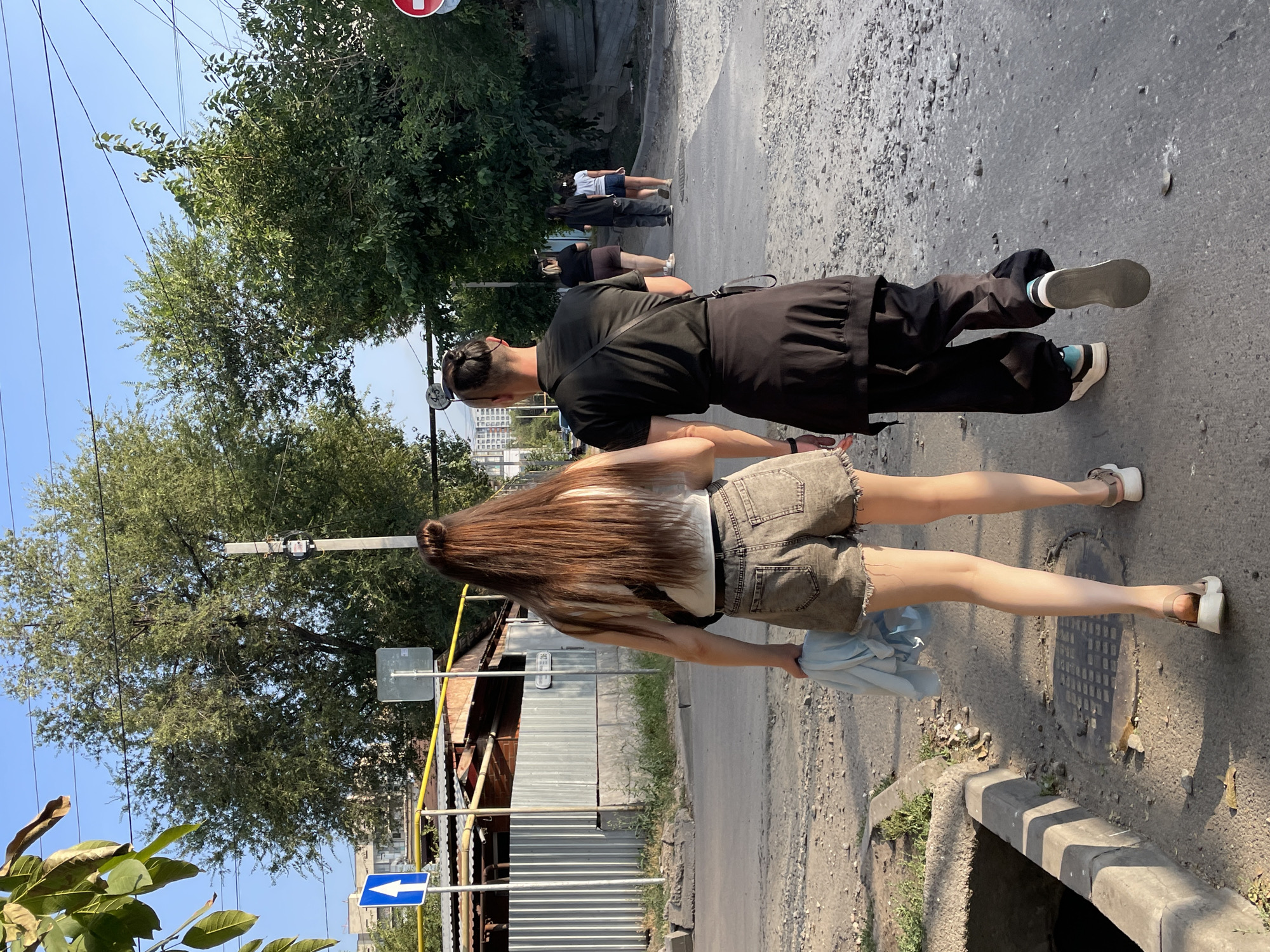 Two individuals walk away from the camera on a gravelly, asphalt-paved path, which appears to be a street or alley in Almaty, Kazakhstan. The person in the foreground on the left is a woman with long, auburn hair tied into a bun. She wears a white tank top, denim shorts, and sandals. She carries a crumpled light blue garment in her left hand. The person to her right is dressed in all black: a t-shirt, baggy pants, and sneakers, with a dark fabric tied around their waist. Their hair is styled in a top knot. Several other individuals are walking ahead of them on the same path, and some buildings, trees, and power lines are visible in the background. The scene is illuminated by sunlight, casting shadows on the ground. A blue traffic sign with a white arrow pointing upward is visible to the left, and a red circular sign is partially visible on the right. The environment suggests a semi-urban or industrial area.
