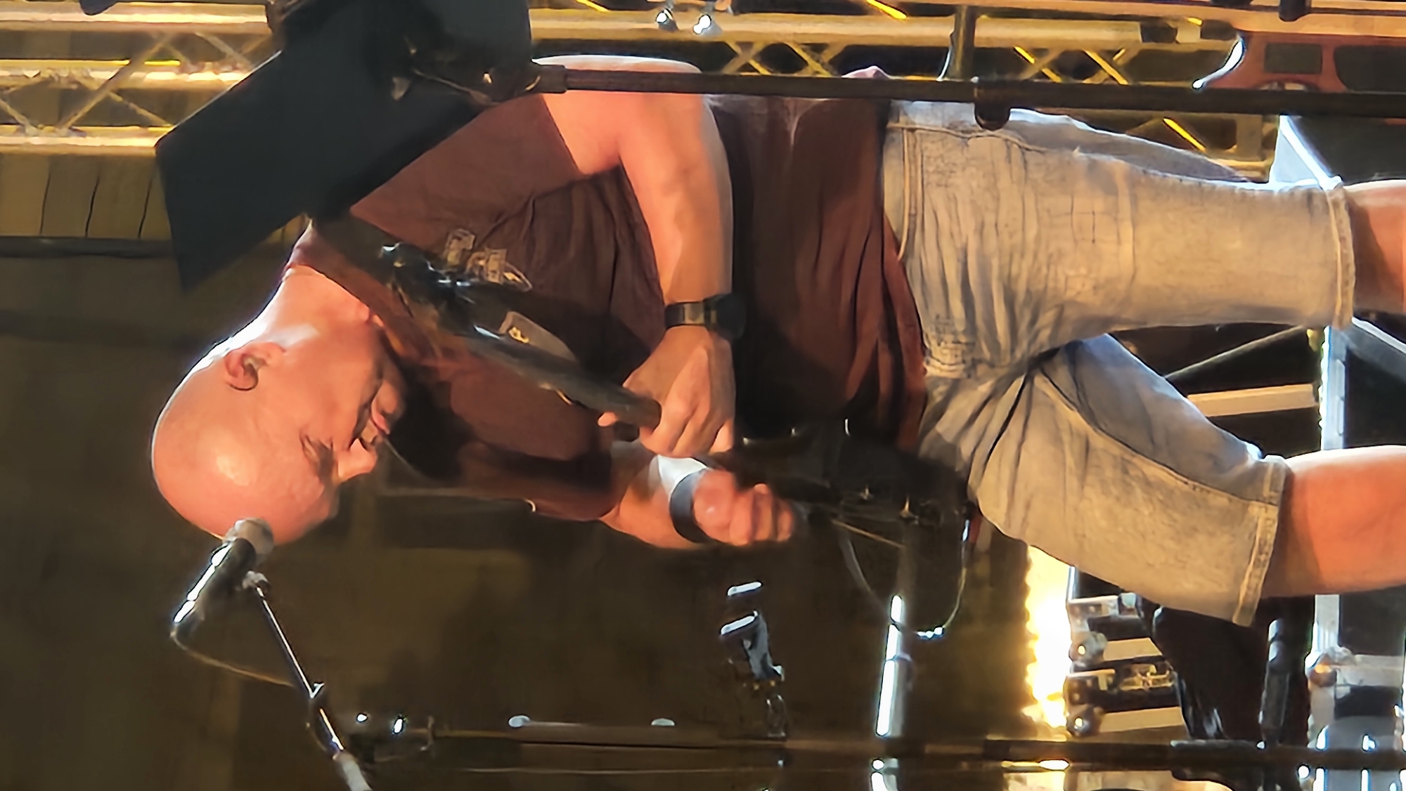 A bald, fair-skinned man with his head tilted down is playing an electric guitar on a stage, possibly during a performance. He is wearing a dark red t-shirt and light blue denim shorts. He has a black wristband on his left wrist. The background is dimly lit, with stage lighting visible and scaffolding structures. The image appears to be captured in the city of Prešov, Slovakia, though the specific venue is not identified.