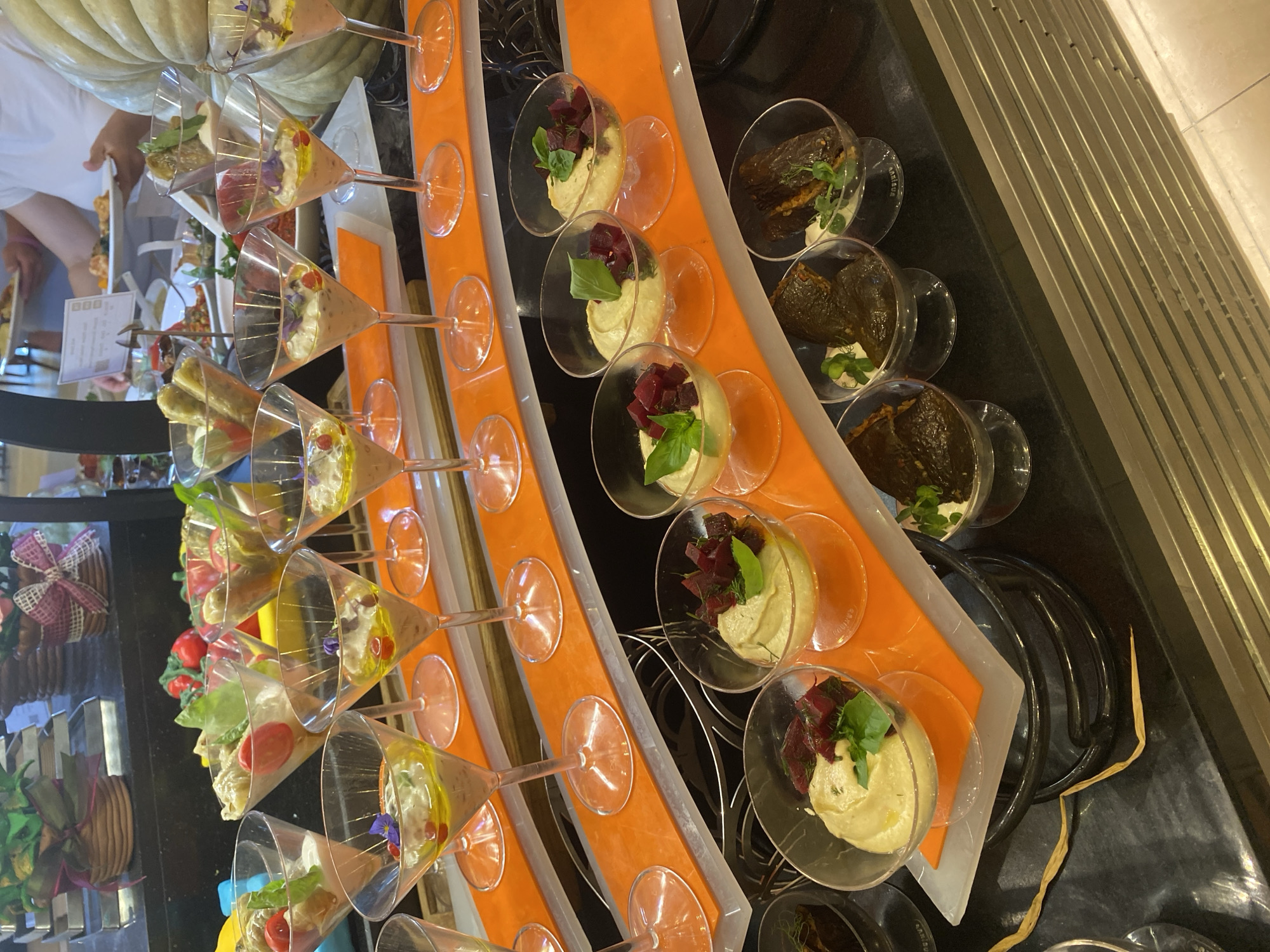 The image shows a close-up view of a buffet table laden with various appetizers, likely at a hotel or restaurant in Aksu, Turkey. The appetizers are predominantly served in individual, clear glass dishes and martini glasses. In the foreground, several martini glasses contain a creamy dip, possibly hummus, topped with herbs, diced beets, and a drizzle of olive oil. Behind these, some martini glasses hold a lighter colored dip with similar garnishes. Further back, various other small dishes are visible, including what appear to be stuffed grape leaves and small rolls filled with a mixture of ingredients, some garnished with green herbs. The buffet table has an orange tiered serving platform. In the background, blurry figures of people can be seen selecting food, with one person holding a white plate. The overall setting suggests a catered event or a buffet spread.