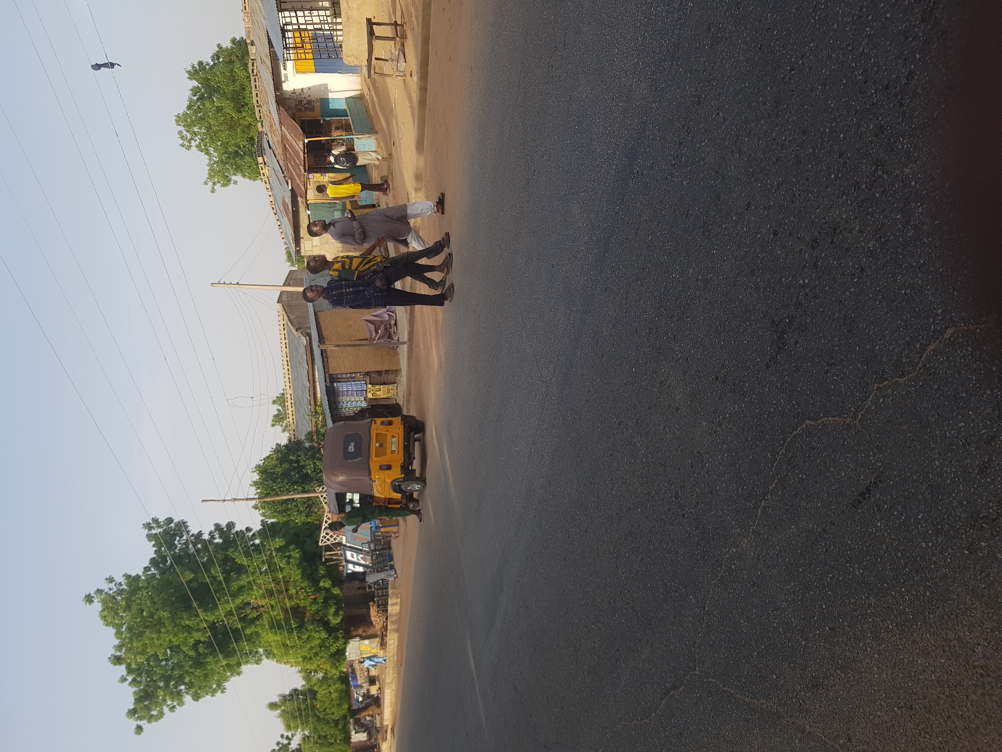 The scene depicts a street in Dabawa, Nigeria. Three men are walking along the edge of the road, alongside a building. A yellow auto-rickshaw is parked nearby. The road itself occupies a significant portion of the image, showing its dark asphalt surface. Buildings, trees, and electrical wires line the roadside, indicating an urban or semi-urban environment.