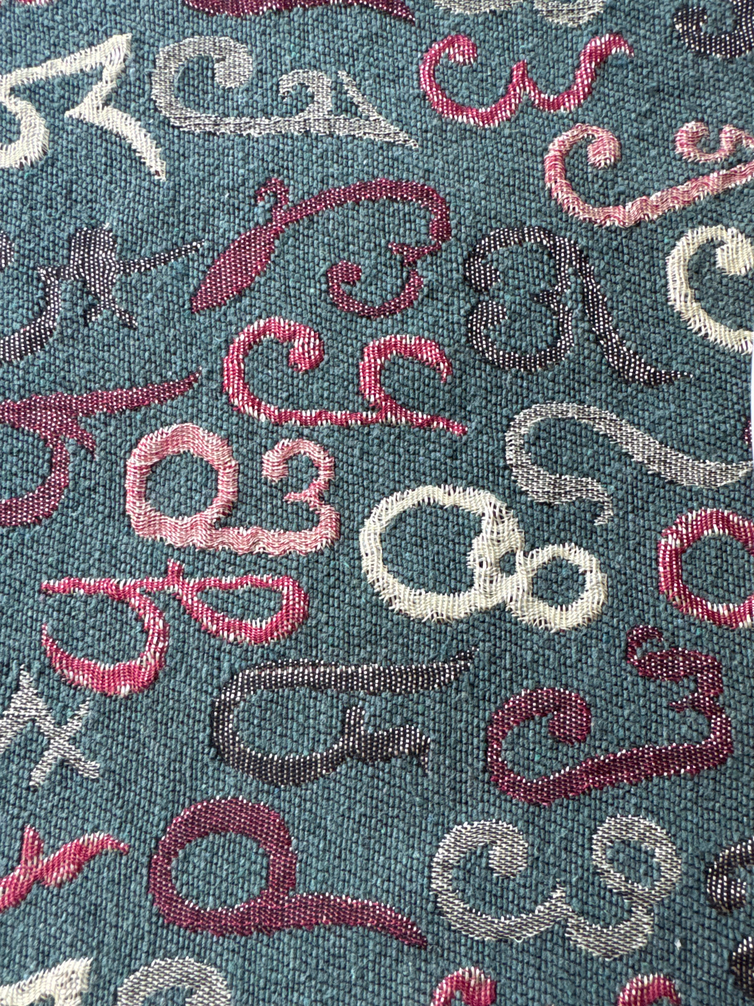 The image shows a close-up of a teal-colored fabric with an embroidered pattern. The pattern consists of various stylized letters and numbers in different colors, including white, pink, burgundy, and black with white speckles. The embroidery has a textured, raised appearance. The specific location in the city of Telavi, Georgia, is not discernible from the image itself. The image is a still photograph and does not depict any actions or interactions.