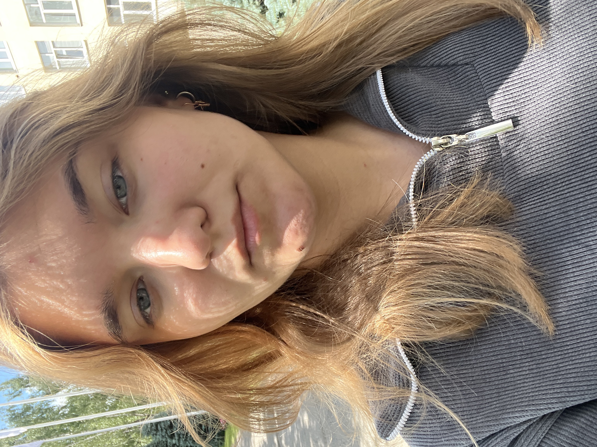 The image is a close-up selfie of a young woman with blonde hair, taken outdoors in Chaikovsky, Russia. She is wearing a grey zip-up hoodie. Sunlight casts shadows on her face, highlighting the texture of her skin and her facial features, including her light blue eyes, light eyebrows, and lips. She has a mole on her cheek and a small blemish on her chin. She is also wearing a hoop earring with a small charm. The background suggests an urban environment with a building with windows and trees visible. The overall lighting indicates it is daytime.