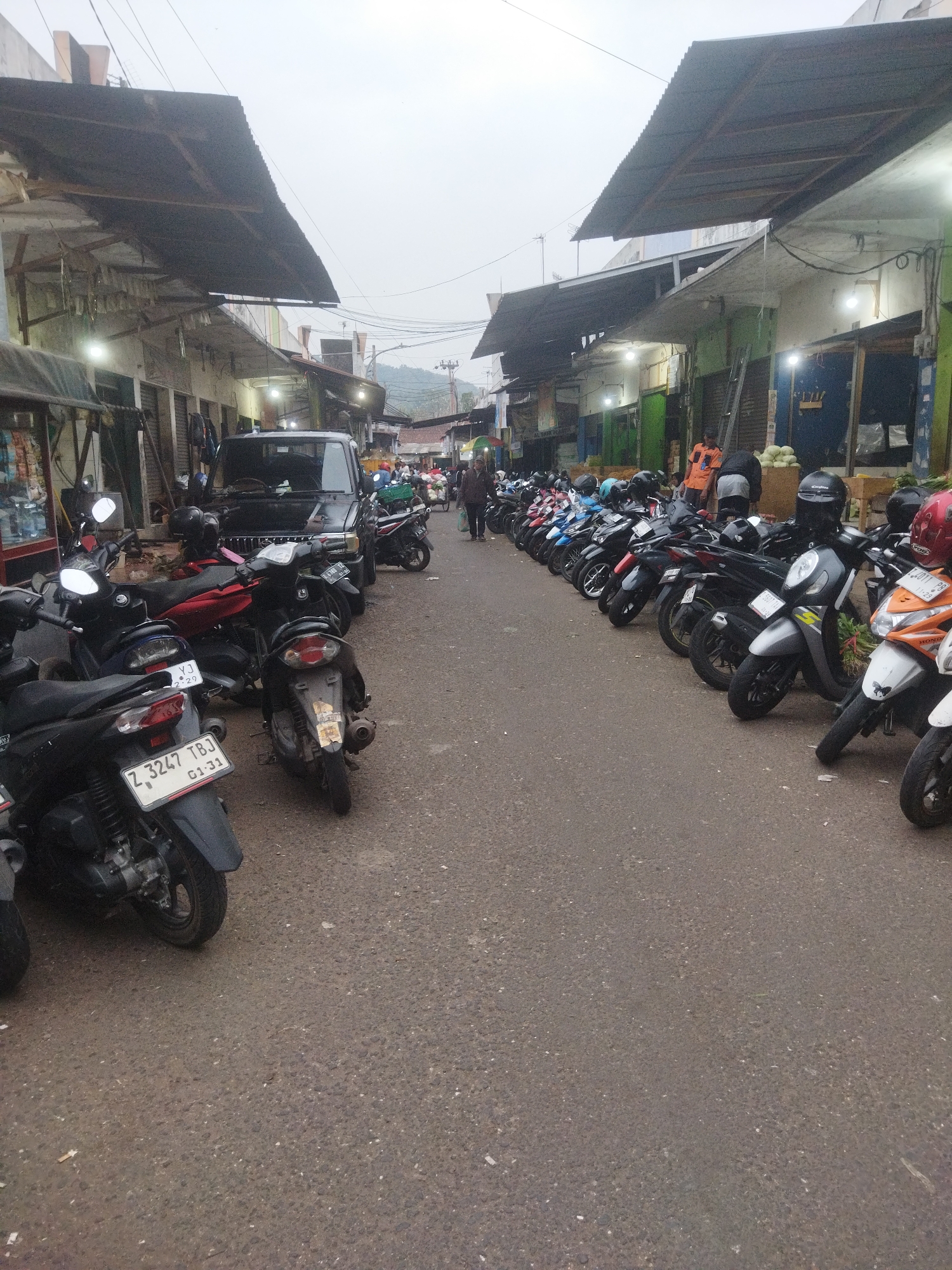 The image depicts a marketplace scene in Banjar, Indonesia, with a focus on parked motorcycles. In the foreground, several scooters are visible, some with prominent license plates such as "Z 3247 TBJ 01-31." The motorcycles are arranged along an asphalt road that runs between rows of market stalls. These stalls have corrugated metal roofs supported by wooden structures and are lined with various goods and shopfronts. People are present, some walking or standing near the stalls, while others are implied to be the owners or users of the parked motorcycles. A black SUV is parked amidst the motorcycles in the mid-ground. The overall environment suggests a bustling but overcast day in a semi-urban market setting.