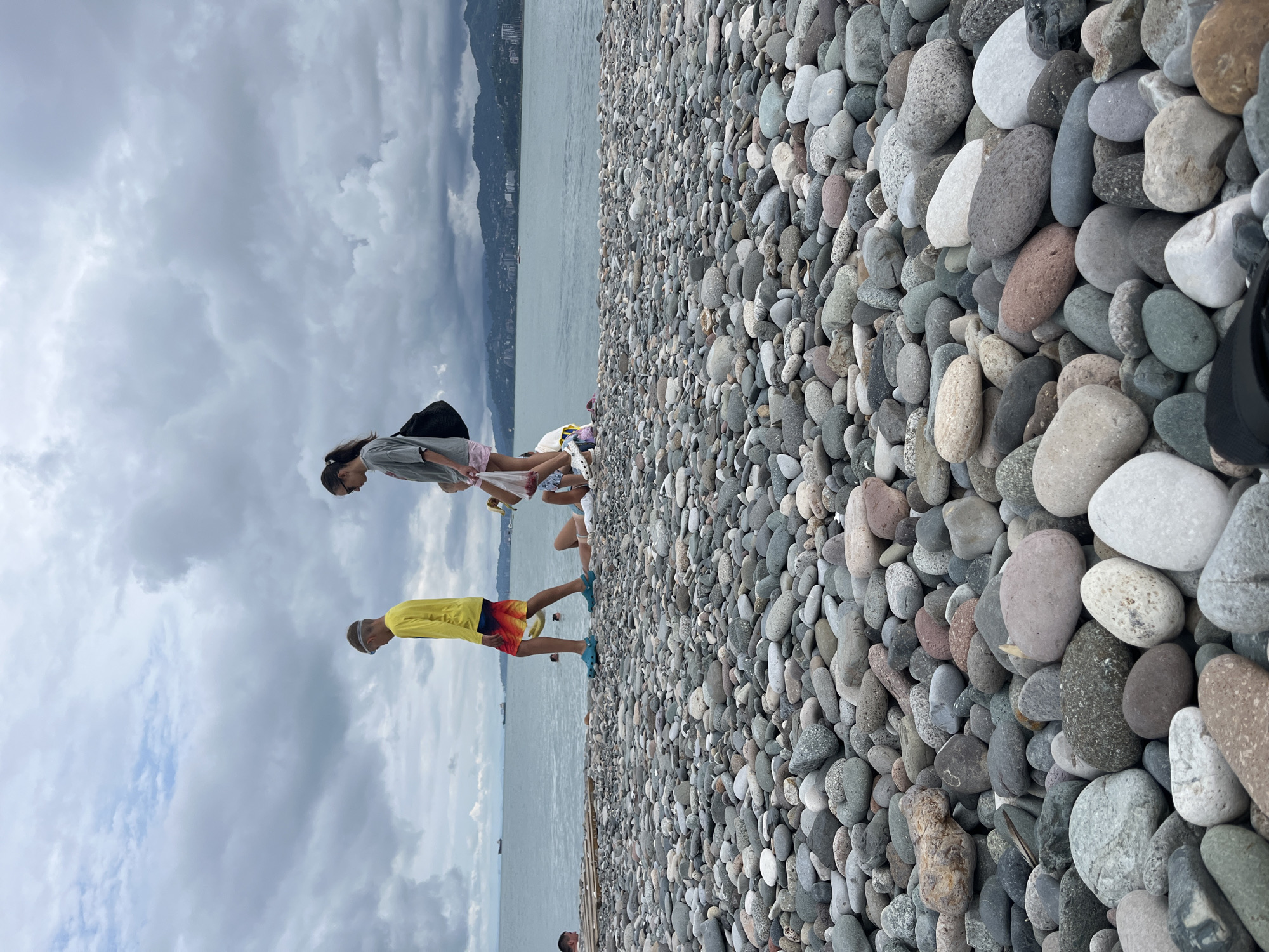 The image depicts a rocky beach in Batumi, Georgia, under a cloudy sky. In the foreground, a vast expanse of smooth, rounded pebbles of various sizes and colors covers the beach. The sea is visible in the background, with a hazy shoreline of distant buildings and mountains. Several people are present on the beach. A boy in a yellow shirt and colorful shorts walks towards the left, wearing blue goggles and blue slides. To his right, a teenage girl in a grey t-shirt and pink shorts carries a dark bag over her shoulder, walking in the same direction. Another child, mostly obscured, is bent over or lying down near the girl. Further to the right, two other children are partially visible, one sitting and the other possibly lying down. The overall atmosphere is that of a casual day at the beach.