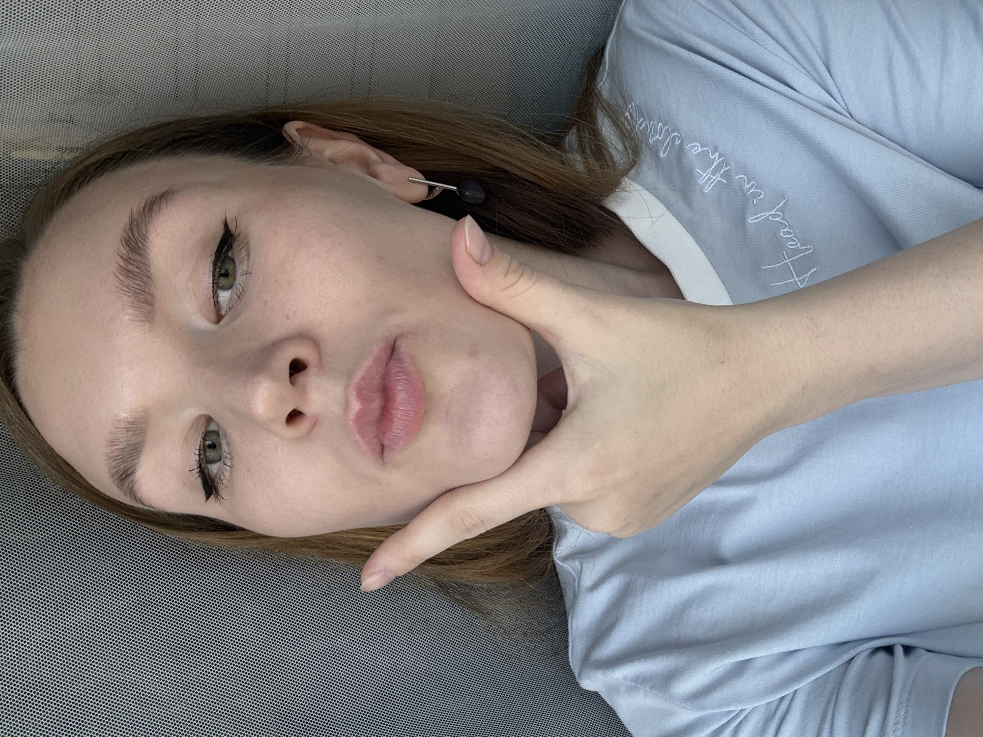 A young woman with light brown hair and green eyes, wearing a light blue t-shirt, poses for a selfie. She is holding her right hand to her chin, with her lips puckered. Her makeup includes eyeliner and mascara. She has a single dark earring in her right ear. The background appears to be a textured grey surface, possibly a wall or screen, and a sheer grey curtain. The text on her t-shirt reads "Head in the clouds" in white embroidery. This image is not geotagged to Izhevsk, Russia.