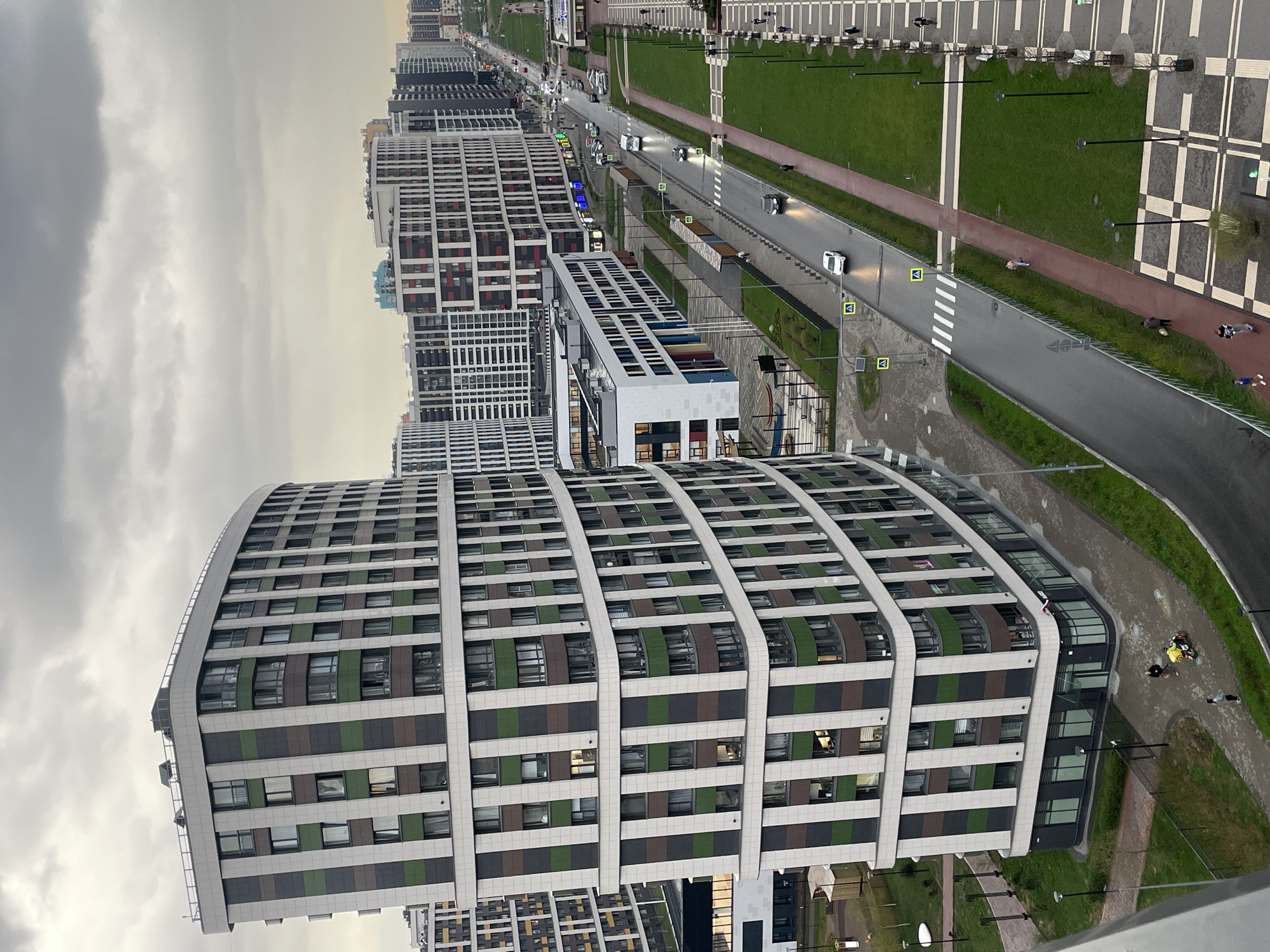 View of apartment buildings and street life in an urban setting in Unknown, Russia. The sky is overcast. In the foreground, there is a curved, multi-story apartment building with alternating green, brown, and white stripes between windows. Other apartment buildings stretch into the distance. A road runs through the scene with cars and pedestrians. Landscaped areas are present on either side of the road. There are parking spots and a pedestrian area at the far right of the image.