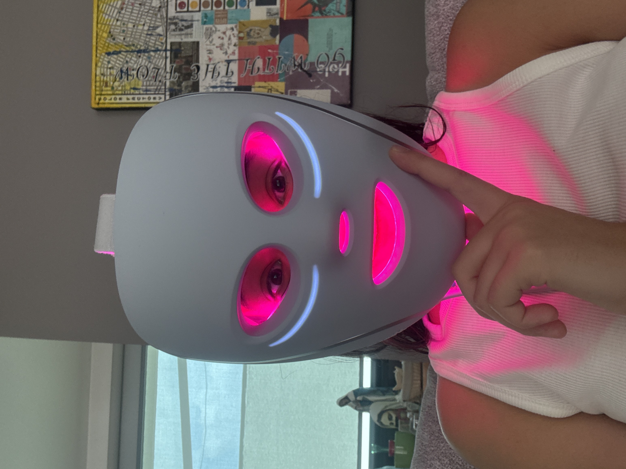 A woman wearing a white LED facial mask is oriented vertically in the image. The mask has openings for the eyes and mouth, with a pink light emanating from these apertures. The mask itself is white. The woman's finger is touching the side of the mask. She is wearing a white tank top. The background includes a grey wall, a colorful artwork, a window, and glimpses of furniture. There is no evidence to suggest that the location is San Bernardino Tlaxcalancingo, Mexico.