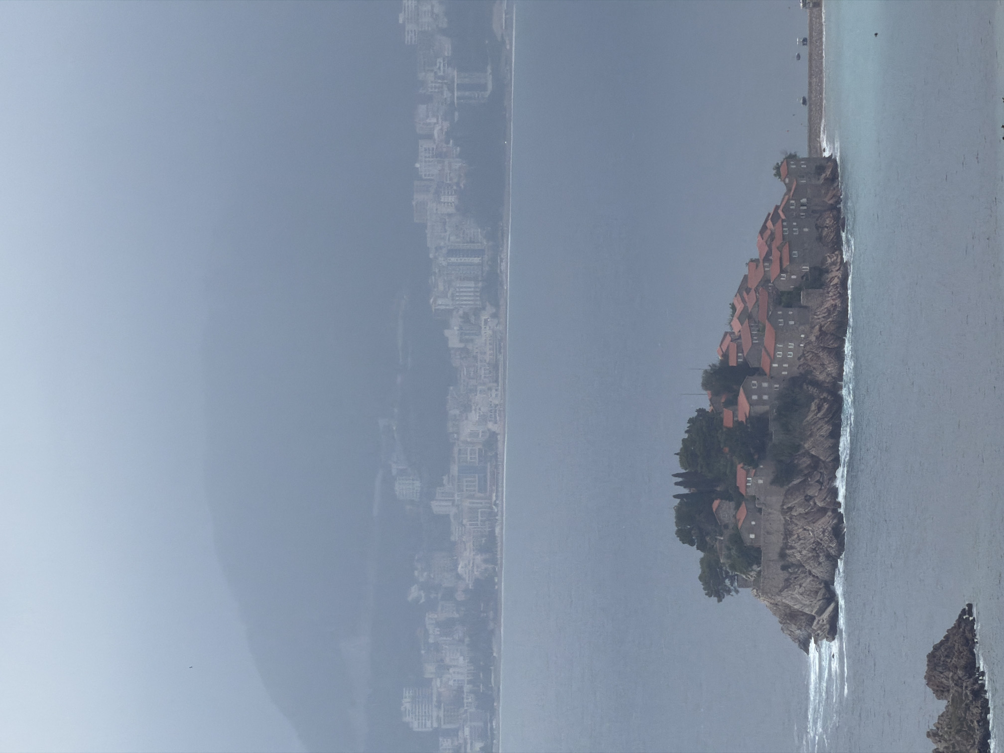 The image captures a panoramic view of a coastal landscape dominated by a distinctive island. On the right side of the frame, a small, rocky island is densely packed with buildings, primarily featuring traditional reddish-orange tiled roofs. Green trees and foliage are visible among the structures and along the island's rocky base. A narrow causeway connects this island to what appears to be the mainland, extending towards the right foreground.
In the far distance, stretching along the left side of the coastline, a sprawling city or town is visible. Its buildings appear lighter in color and are somewhat obscured by haze or distance, blending with the atmospheric conditions. The middle and foreground are filled by a large expanse of calm, bluish-grey water, likely the sea, with gentle waves noticeable around the island and along the shore. A few smaller rocky formations are also present in the water in the immediate foreground. The sky above is a uniform, soft light grey-blue, suggesting an overcast or hazy day.
No specific human activity is depicted; the scene is a serene landscape. The muted colors and soft lighting indicate daytime under hazy or cloudy weather conditions. The iconic appearance of the island village, connected by a causeway to the mainland, clearly identifies the location as Sveti Stefan in Montenegro. There is no visible text in the image. The overall impression is one of tranquility and historical charm.