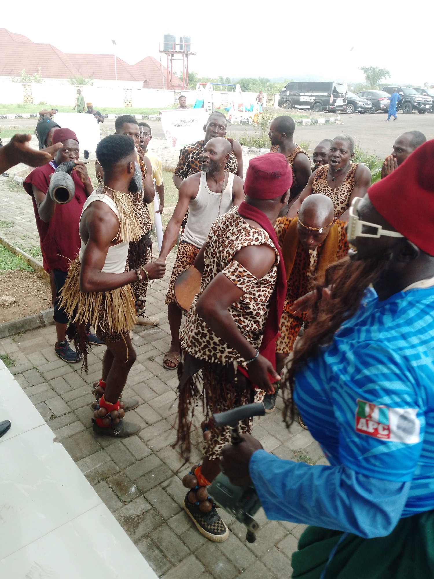 In an outdoor setting on a paved surface, a group of people are engaged in a cultural performance or celebration. Most of the individuals are dressed in traditional clothing, some featuring animal print patterns and grass skirts. Some are playing musical instruments such as drums and horns. Several individuals are wearing what appears to be leg rattles or ankle bells. The background includes buildings, parked vehicles, and trees, suggesting the location might be a public space or event venue. One individual wears a blue shirt with the acronym "APC" stitched on the sleeve. Location is identified as Unknown, Unknown.