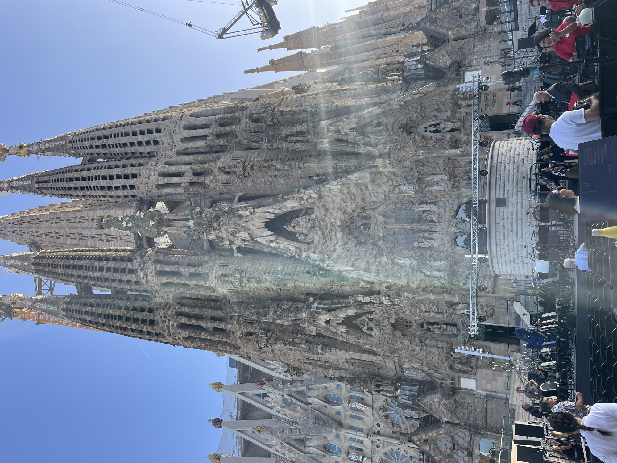 This image depicts a low-angle view of the Sagrada Familia in Barcelona, Spain, on a clear, sunny day. The iconic basilica dominates the upper portion of the frame, with its intricate Gothic and Art Nouveau architecture, including multiple spires and detailed facade carvings, visible against a bright blue sky. Construction cranes are present, indicating ongoing work on the basilica.
In the foreground, a stage area has been set up, with rows of empty chairs for an audience. Various stage equipment, including speakers, lighting rigs, musical instruments (such as a cello in a case), and microphones, are arranged on and around the stage. Several people are visible in the foreground and around the stage area, some appearing to be staff or performers. They are casually dressed and engaged in various activities, such as setting up equipment or conversing. The overall scene suggests a public event or performance taking place in front of the Sagrada Familia.