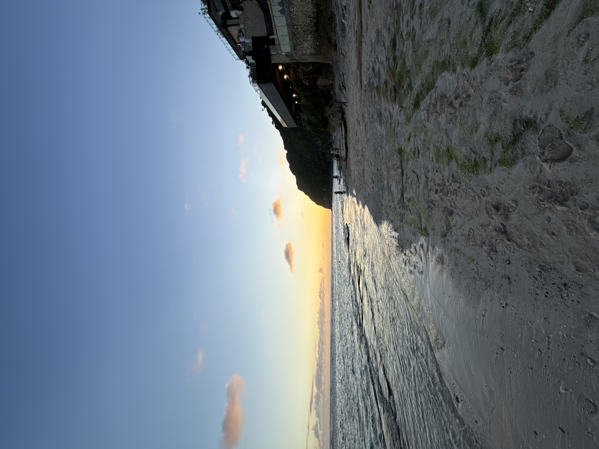 The image shows a view of a beach at sunset in Ungasan, Indonesia. The sky transitions from blue to orange near the horizon, with scattered clouds. A cliff covered in greenery and topped with a multi-story building is visible along the beach. People are walking along the shoreline. The beach is sandy, and the water is calm.