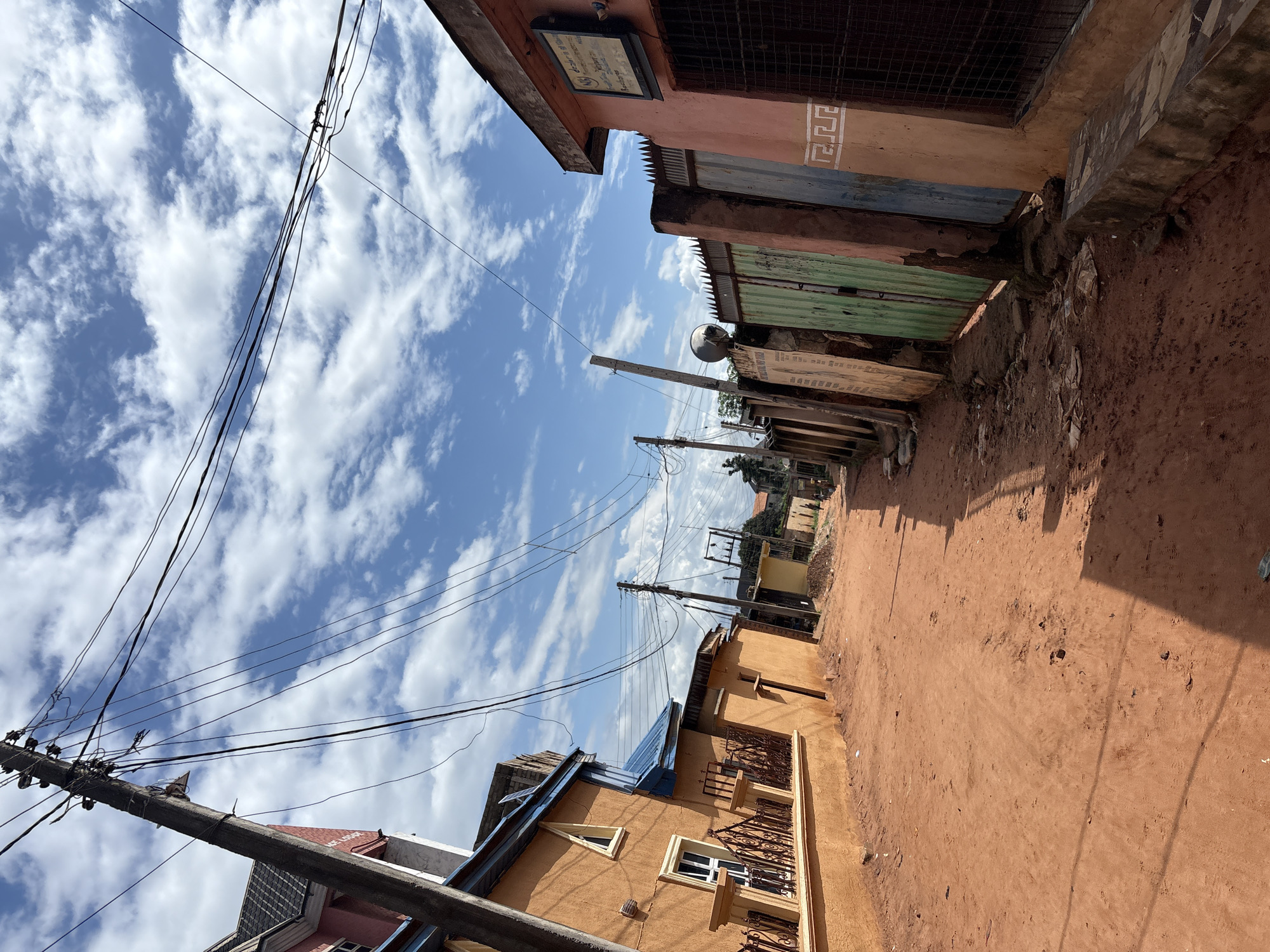 The image depicts a narrow, unpaved street in Nkpor, Nigeria, characterized by red earth and sparse vegetation. On the left side of the street, an orange building with blue roofing is visible, featuring decorative fencing and windows. A wooden utility pole with numerous electrical wires is prominently positioned in the foreground, leaning to the left. The right side of the street features a row of buildings with corrugated metal gates in various colors, including green and blue. A pink building with a sign above its entrance is also present. The sky is partly cloudy with a bright blue sky. The scene suggests a residential or mixed-use area within a Nigerian city.