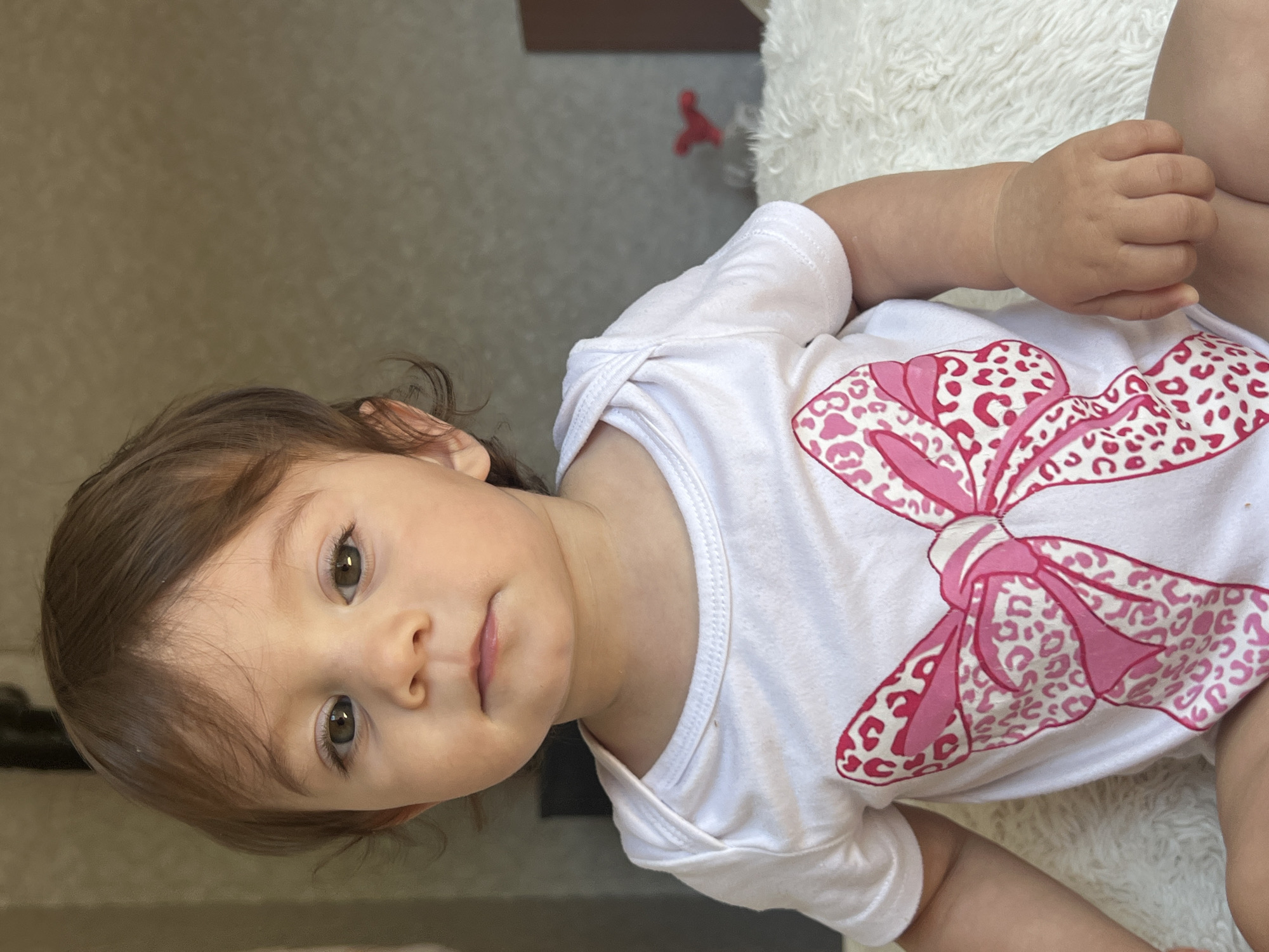 A close-up shot shows a young child, likely a toddler, seated in a fluffy white rug or blanket. The child has brown hair, light skin, and brown eyes, looking directly at the camera with a neutral expression. They are wearing a white bodysuit with a large pink bow pattern that features a leopard print design. The background is blurred, with a neutral wall and hints of furniture, suggesting an indoor setting, possibly a bedroom. The image quality is good, with natural lighting. No specific location in Elena, Bulgaria can be identified from the image itself.