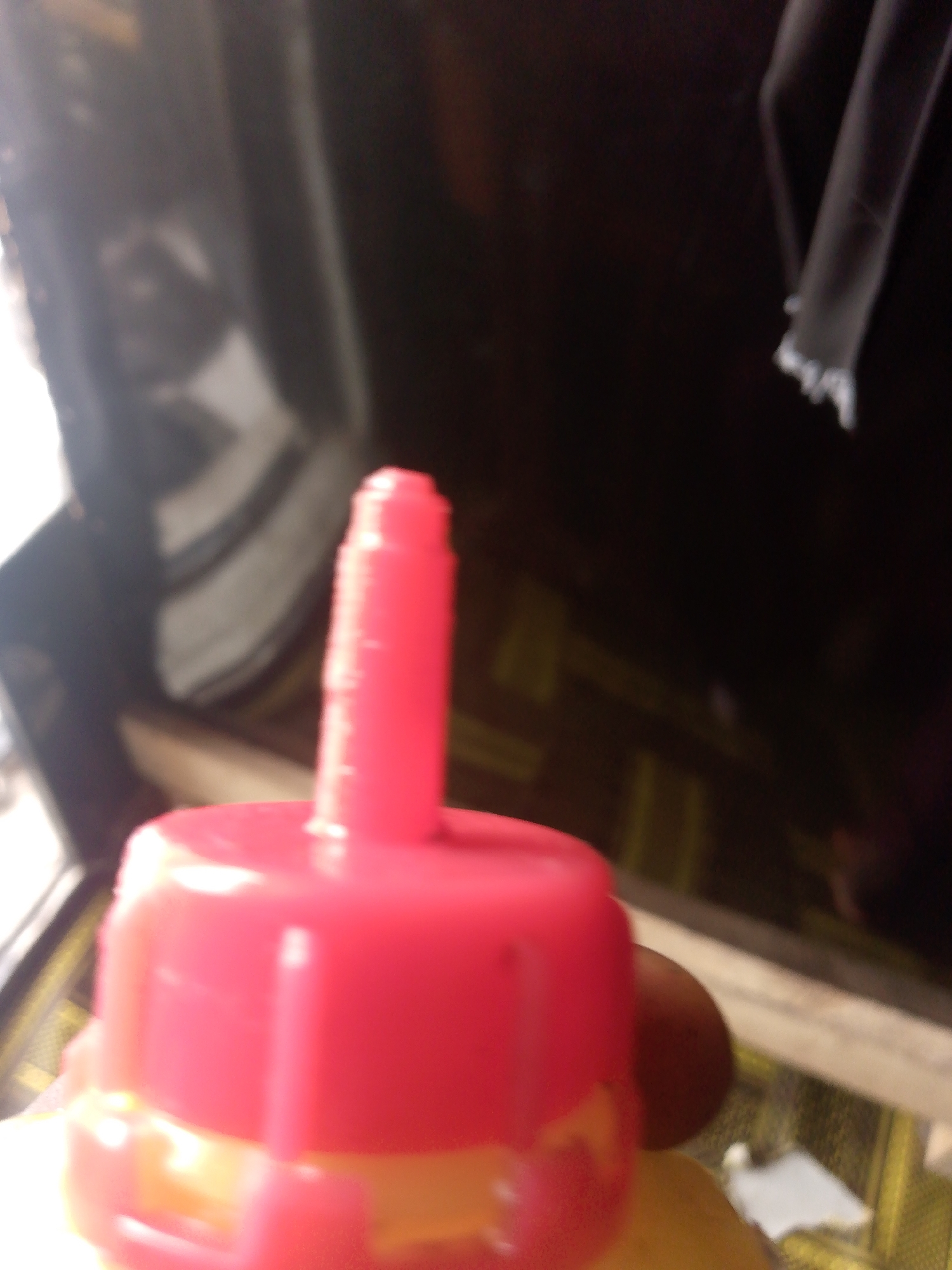 The image shows a close-up of a red bottle cap with a nozzle. The cap is hexagonal and has a cylindrical nozzle extending upwards. The background is blurred, but appears to be an indoor setting with some reflective surfaces and a dark color scheme. The lighting is bright, casting shadows and creating highlights on the red plastic. There is no discernible location in the city of Jauro Yinu, Nigeria.