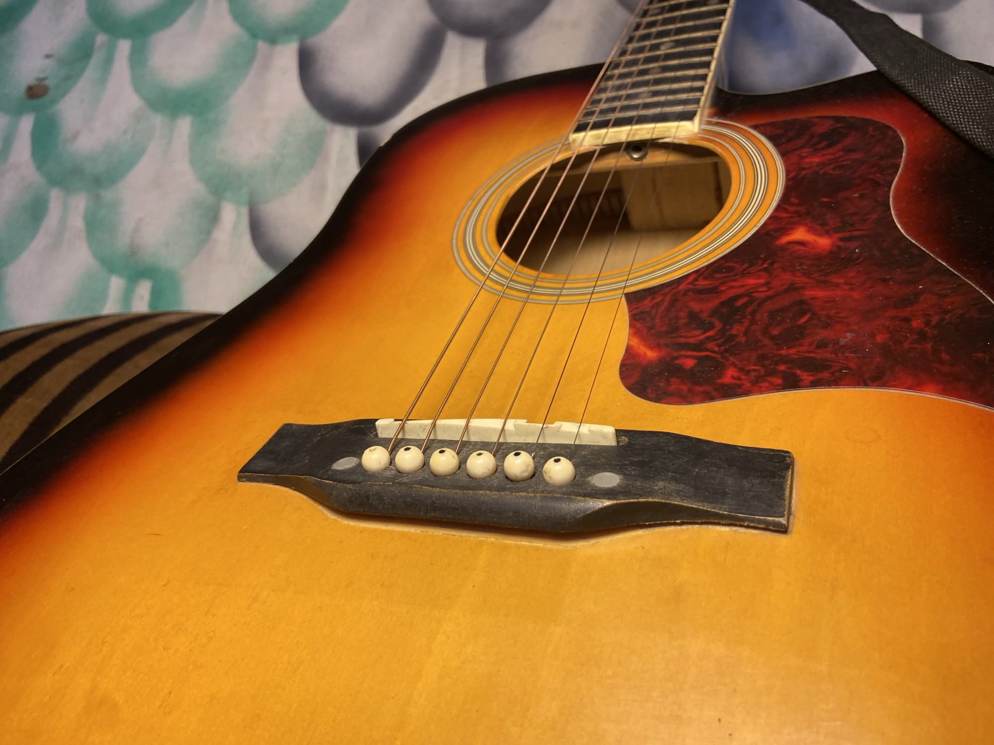 A close-up, low-angle shot captures the body of an acoustic guitar. The guitar displays a classic sunburst finish, transitioning from a warm yellow-orange in the center to a dark red-brown towards its edges. Six steel strings extend over a dark wooden bridge, which holds a white saddle and six off-white bridge pins. A reddish-brown, tortoiseshell-patterned pickguard is affixed to the right of the circular soundhole, which is framed by decorative rings. The lower portion of the guitar's fretboard is visible towards the top-left, extending from the neck. A black fabric strap is draped over the upper right side of the guitar.
The background behind the instrument features a light-colored surface with a repeating pattern of light blue-green, scalloped or scale-like shapes. Some darker, indistinct circular shapes are also present in the background. In the bottom-left corner, a striped object with alternating dark and light brown bands, possibly a cushion or part of a chair, is partially visible. The guitar shows minor signs of wear, including dust on its surface and bridge. No people are present, and no activity is depicted. The lighting indicates an indoor setting. No text is visible in the image.