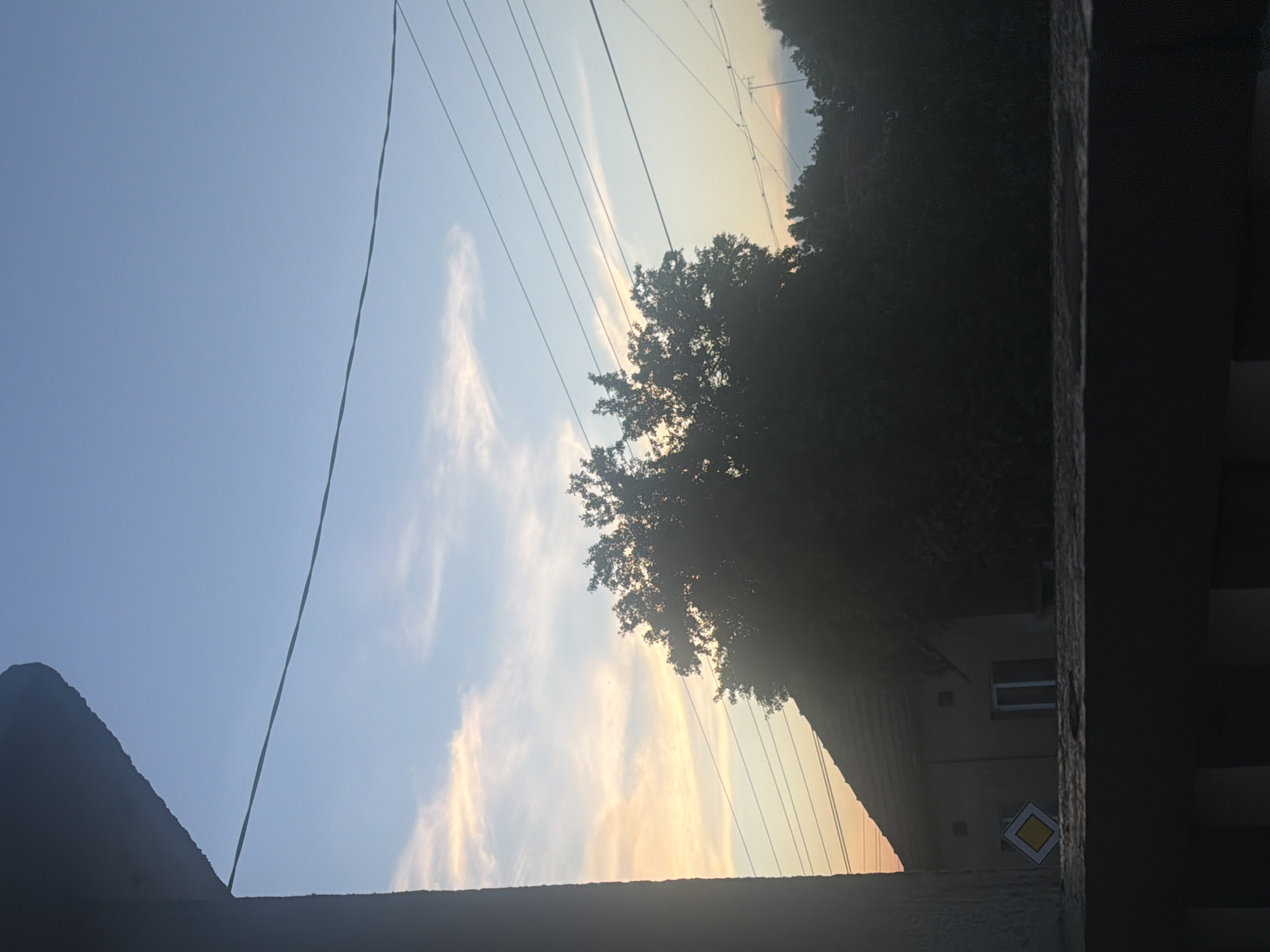 The image displays an outdoor scene captured during dusk or dawn, indicated by the sky's color gradient. The upper portion of the frame is dominated by the sky, which transitions from a clear, light blue on the left to warm orange and yellow tones on the right. Sparse, wispy clouds are scattered across the sky.
Several thin power lines or utility wires stretch diagonally across the upper and middle sections of the sky. A large, dense tree is silhouetted prominently against the lighter part of the sky on the right side of the image. Below the tree line, parts of buildings are visible, largely in shadow. One building shows a single, rectangular window with a white frame.
In the lower right quadrant, attached to a building or wall, a distinct yellow diamond-shaped road sign with a white border and a black outline is visible. This type of sign typically indicates a "priority road" in European traffic regulations. The foreground features dark, silhouetted architectural elements: a sloped roofline on the bottom left and a textured, dark wall or pillar framing the entire right side of the image, suggesting the photograph was taken from a balcony or window. No people or specific activities are visible, conveying a quiet, contemplative atmosphere in what appears to be a residential or semi-rural setting.