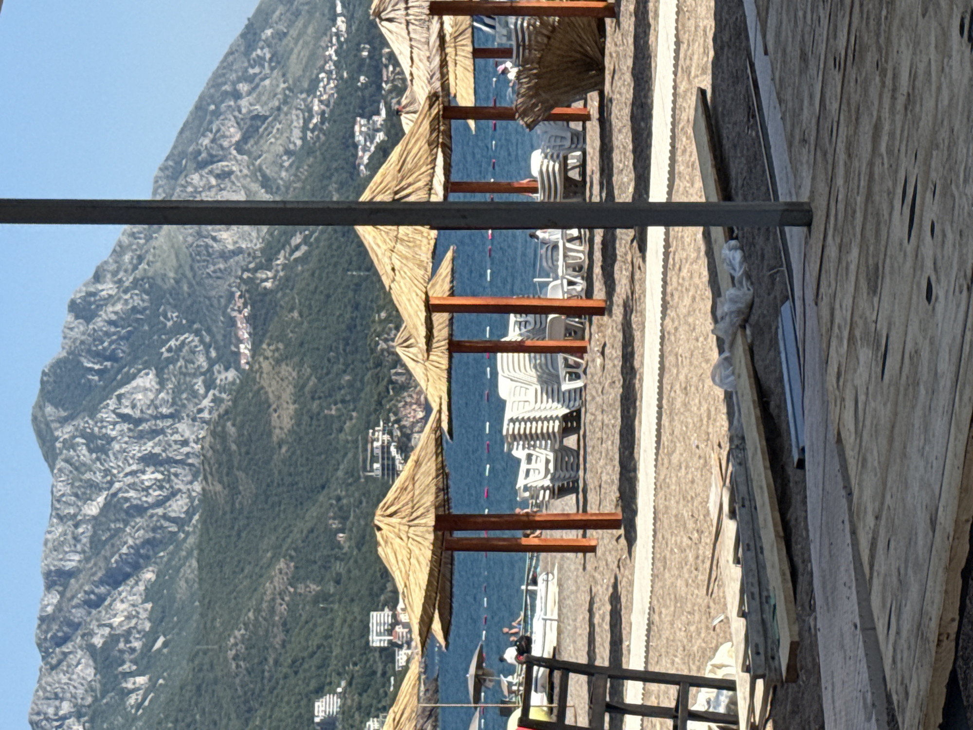 This media file depicts a beach scene in Boreti, Montenegro. The foreground features a wooden walkway or deck under construction, with raw wooden planks and some construction materials scattered around. Beyond the walkway, a sandy beach is visible, lined with rows of straw-thatched umbrellas supported by wooden poles. Stacked white beach chairs are neatly arranged under the umbrellas, suggesting a resort or public beach area.
The middle ground of the image is dominated by the deep blue sea, with a few buoys marking an area on the water. A person can be seen partially obscured by the beach umbrellas, sitting on a white beach chair and looking towards the water. In the background, a large, majestic mountain rises steeply, its rocky, textured surface covered in patches of green vegetation. Along the lower slopes of the mountain and extending towards the sea, a town or village with white buildings is visible, nestled among the trees.
The sky above is a clear, bright blue, indicating fair weather and a sunny day, likely midday or afternoon given the lighting and shadows. The overall impression is one of a serene coastal landscape, possibly during the peak tourist season, with elements of both natural beauty and human development. There is no visible text in the image.