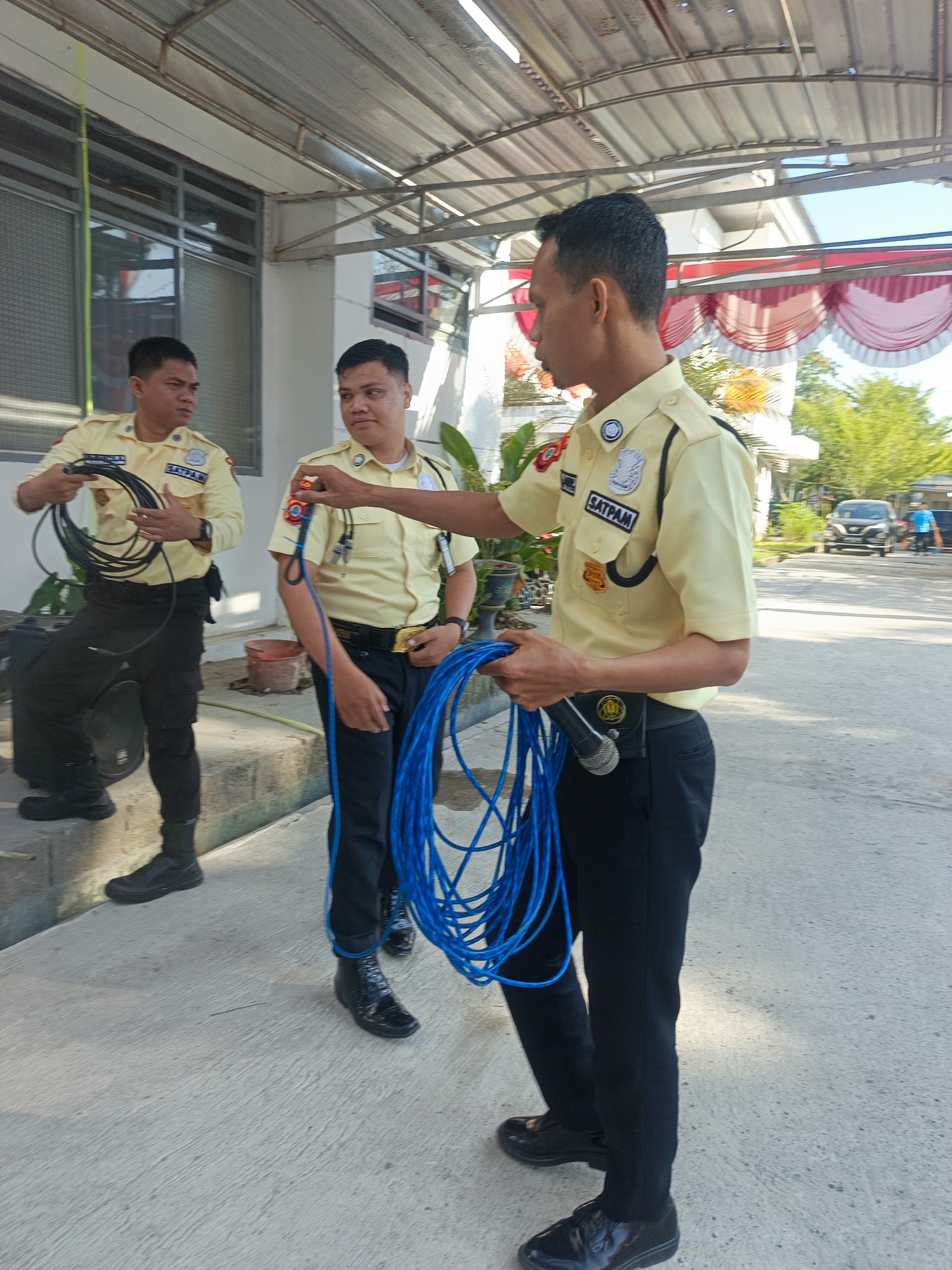Three male security guards, dressed in light yellow uniforms and black pants, are standing outdoors in what appears to be the grounds of a building. The guard on the right is holding a coil of blue cable and a microphone attached to his belt. The guard in the middle is holding a coil of black cable. The guard on the left is also holding a coil of black cable. The building has a corrugated metal roof supported by white beams. A red and white draped fabric is visible on the building. In the background, there is a road with a dark-colored car and a person in a blue shirt walking. Trees and foliage are also present in the background, suggesting an outdoor environment in a tropical or subtropical region. The location is specified as the city of Gorontalo, Indonesia.