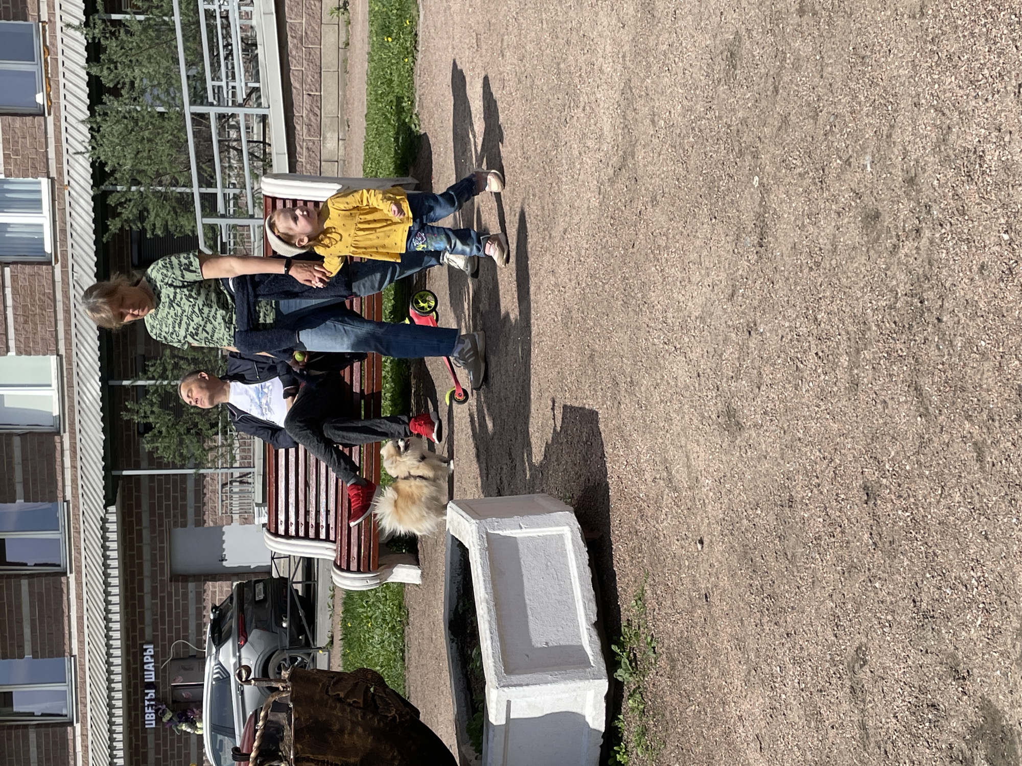 The image depicts a family scene in Saint Petersburg, Russia. An adult woman with blonde hair and wearing a green patterned shirt sits on a park bench alongside a young child in a yellow dress and jeans. The woman is holding the child's hand. A second adult male, wearing a white t-shirt and dark jacket, also sits on the bench. A small white fluffy dog stands on the ground near the bench. A red scooter is near the child.
The scene takes place outdoors on a gravelly surface. A white rectangular planter is visible near the bench. In the background is a brick building with windows and a metal canopy over the entrance. A car is partially visible behind the bench. A sign with text in Russian, "ЦВЕТЫ ШАРЫ" (Flowers, Balls), is present. The weather appears to be sunny, casting shadows of the people and objects on the ground.