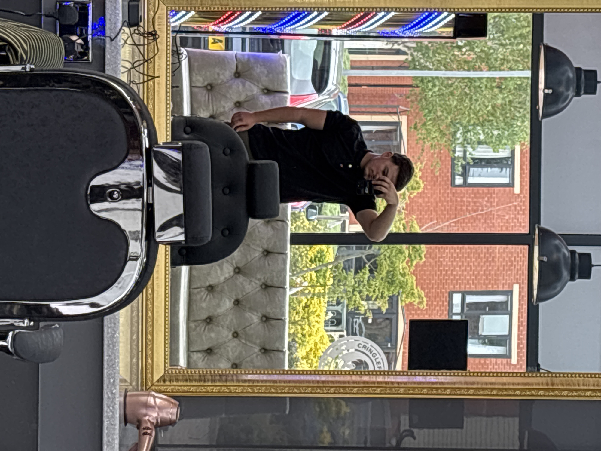 An interior shot shows a barber's mirror reflecting a customer in a black shirt, seated on a tufted seat. The reflection shows an exterior view of a street with a red brick building, trees, and cars, including a white vehicle with visible license plates. The mirror's frame has an ornate gold finish and is decorated with a barber pole pattern along the top. In the foreground, part of a barber's chair is visible, along with a hair dryer. A storefront with the name "CRINGLEFORD" is seen in the reflection. Based on this storefront, the location is likely Cringleford in Norwich, United Kingdom.