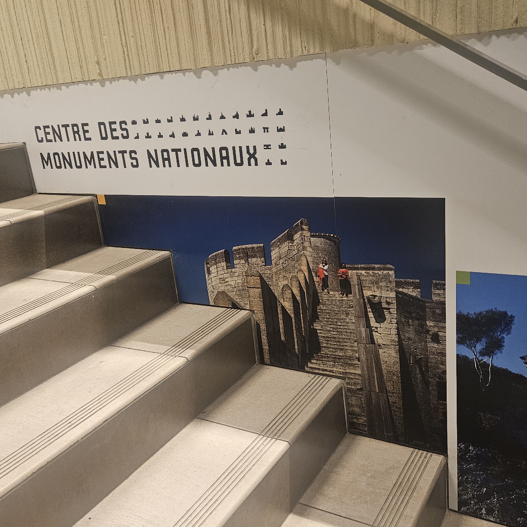 The image shows a close-up view of a staircase with a large banner displayed on the wall above it. The banner features text that reads "CENTRE DES MONUMENTS NATIONAUX" with small silhouetted images of buildings. Below the text, there is a photograph of a stone castle structure with people on its steps. The staircase is made of light-colored stone and has textured lines on each step for grip. The environment appears to be indoors, possibly a museum or public building, given the banner and the staircase design. The castle photograph on the banner is a prominent feature, depicting a historical architectural site. The image does not provide explicit details about the city of Lançon-Provence, France, but the banner suggests a connection to national monuments.