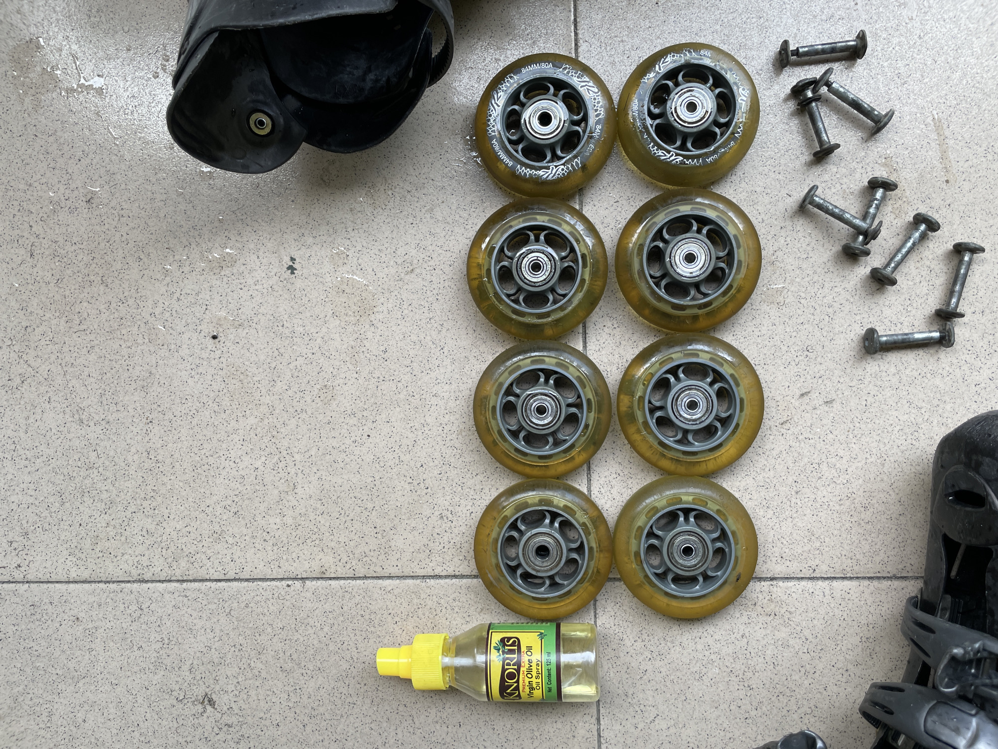 The image depicts a collection of inline skate wheels and associated hardware, along with a bottle of "Knoris Virgin Olive Oil Oil Spray," arranged on a tiled surface. There are eight yellowed, translucent inline skate wheels, each with a grey inner hub and bearings. Several screws and bolts are scattered around the wheels. In the upper right corner, a black plastic part of an inline skate is visible. The tiled floor appears to be outdoors, with some light water spatter visible on the surface. The scene is static and shows no people or ongoing actions. The location is Ado, Nigeria.