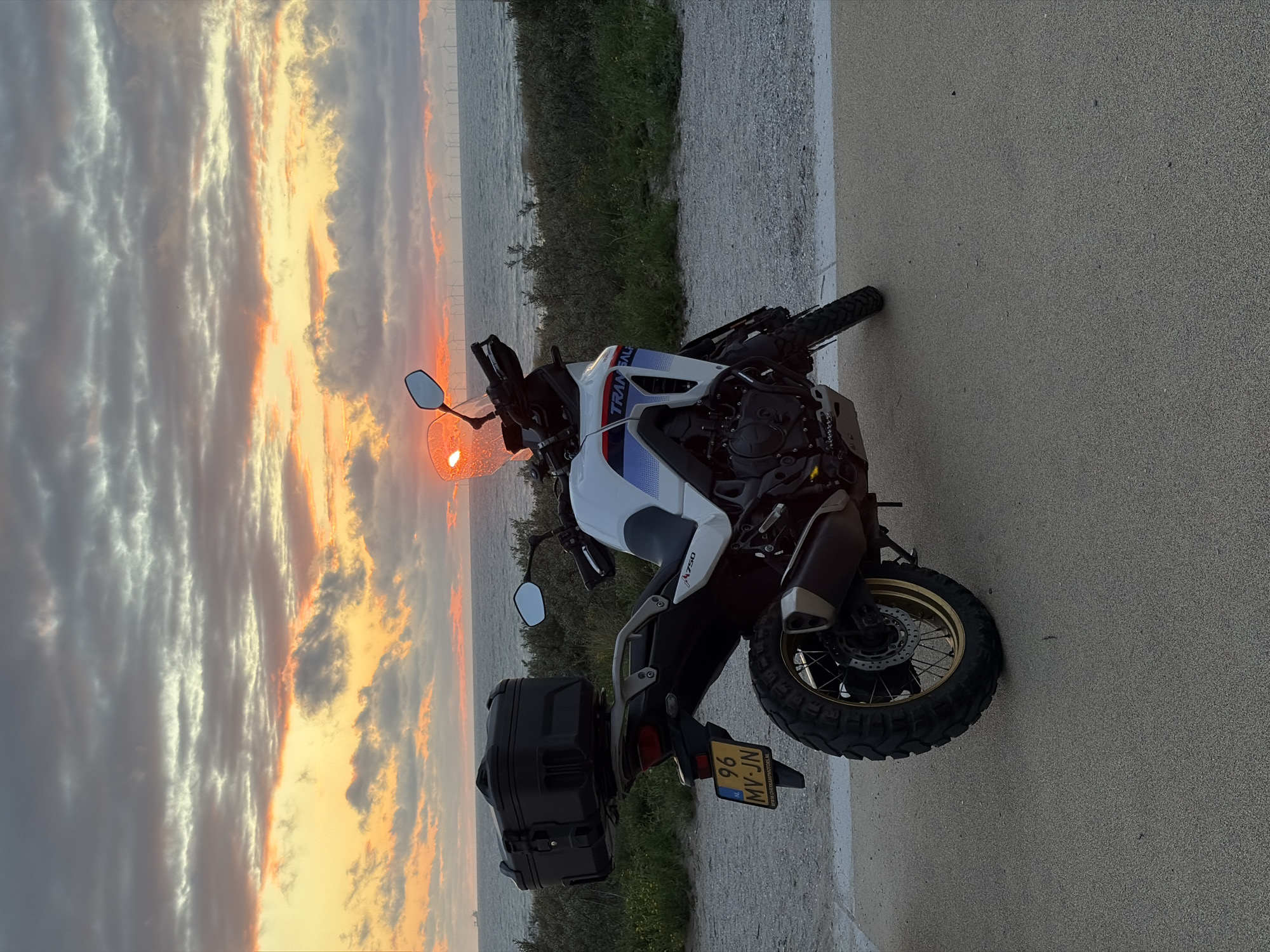 A white and blue motorcycle is parked on a paved surface facing right. It has a black storage box mounted on the rear rack and a license plate that reads "96 MV-JN". The motorcycle is positioned in front of a body of water, likely the sea, under a dramatic sunset sky with clouds in hues of orange, yellow, and gray. In the distance, a row of windmills is visible on the horizon. The foreground shows a gravel path bordering the paved area. The scene appears to be a coastal location in the city of The Hague, Netherlands, given the Dutch license plate and the characteristic Dutch landscape with windmills. There are no people visible in the image.