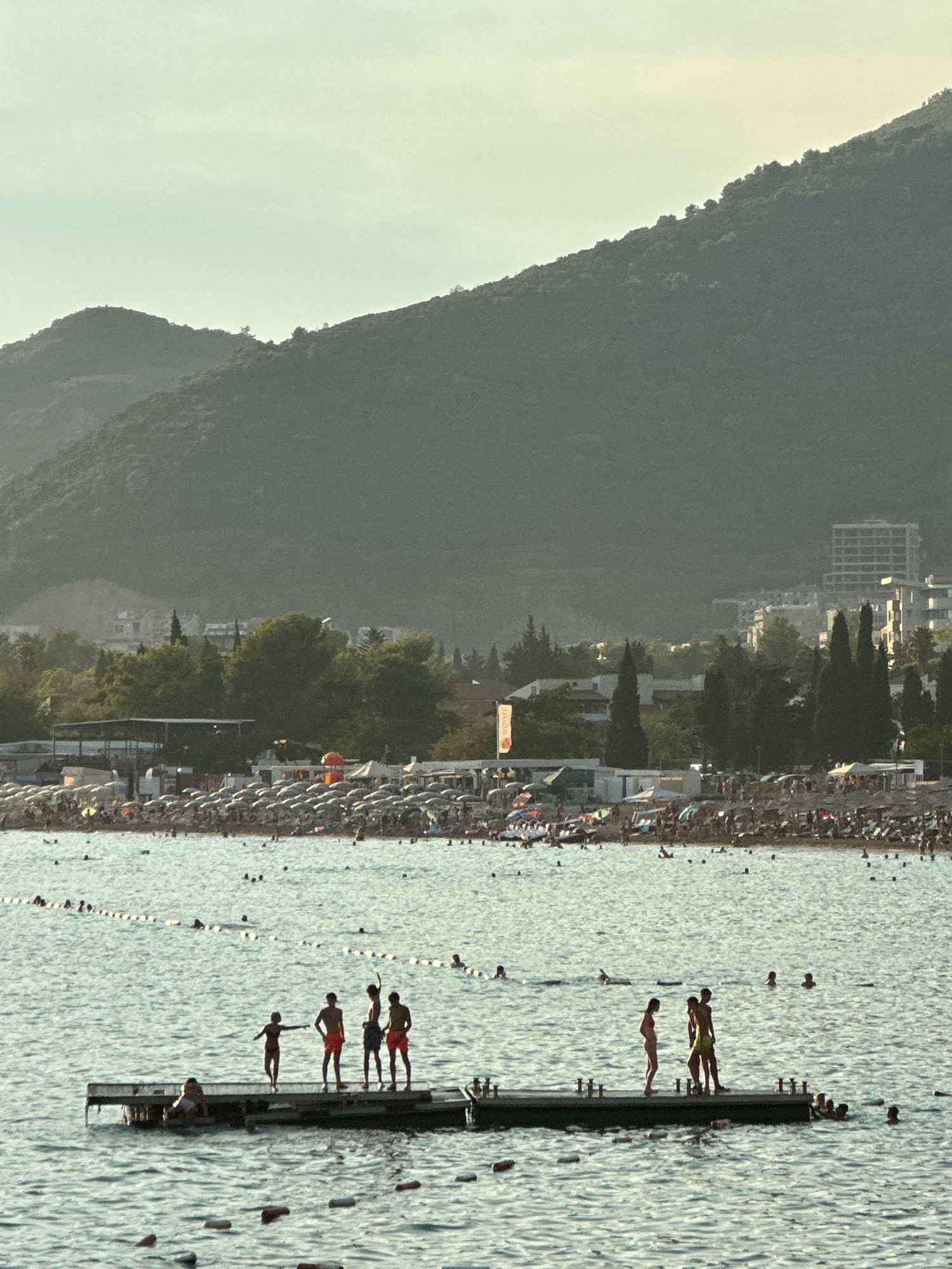 In Budva, Montenegro, a group of young people are gathered on a floating wooden platform in the sea. Several individuals are standing, some appear to be talking or gesturing, while others are seated or lying down. The platform is positioned in front of a crowded beach lined with numerous umbrellas and people. Beyond the beach, trees and buildings, including a multi-story structure, are visible, backed by a large, tree-covered mountain that dominates the background. The sky above is hazy, and the overall lighting suggests it is either early morning or late afternoon.