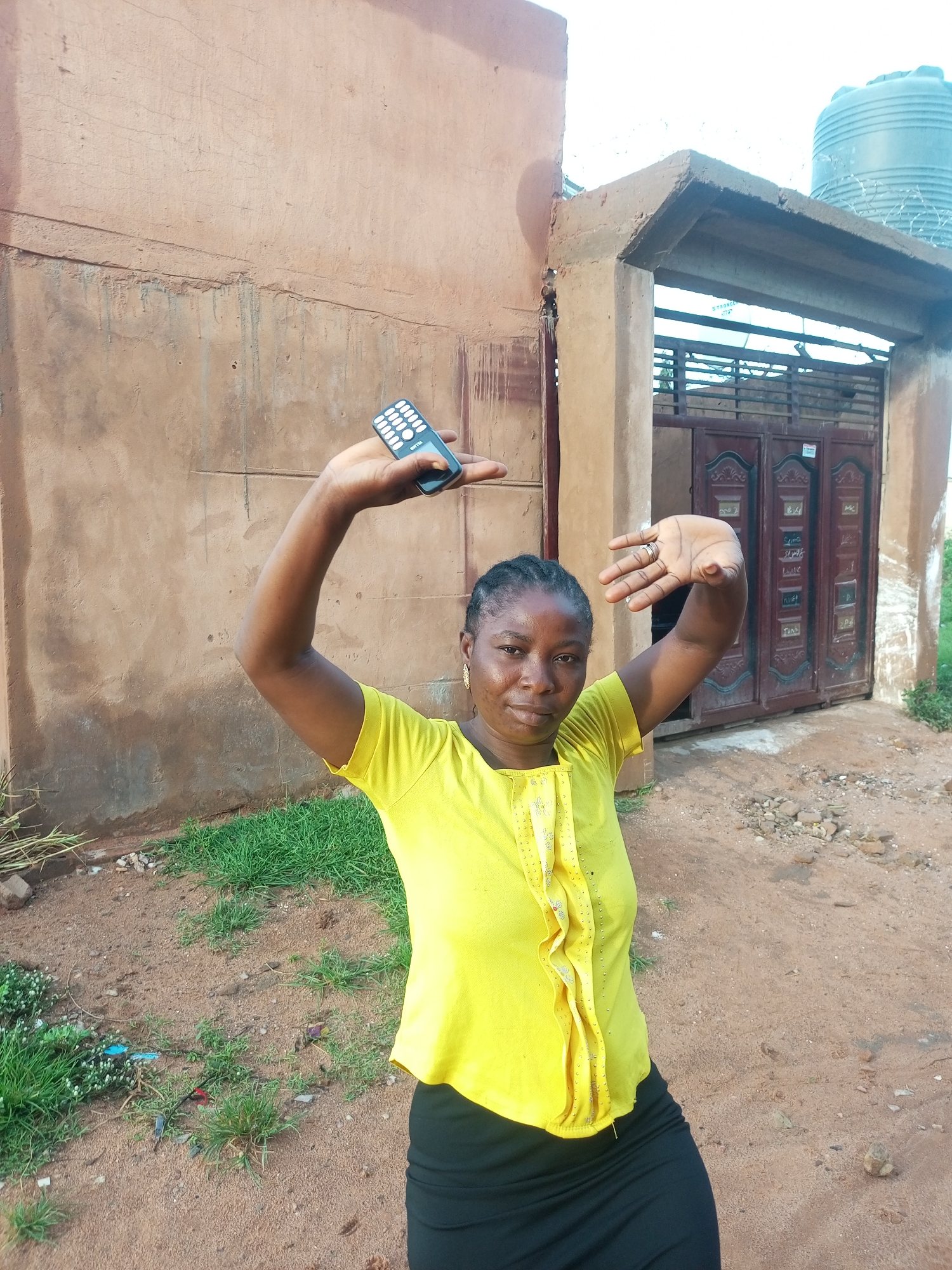 A woman stands outside a building in Tempure, Nigeria, holding a black mobile phone in her right hand, extended above her head. Her left arm is also raised, palm open, in a defensive or signaling gesture. She wears a bright yellow t-shirt with a ruffled, decorative front and a black skirt. The building behind her is constructed of brown, textured walls. To the right, a large metal gate with decorative panels is visible, and above it, a large plastic water tank sits on the roof of the structure. The ground is covered in reddish-brown dirt with scattered rocks and patches of green grass. The sky is overcast.