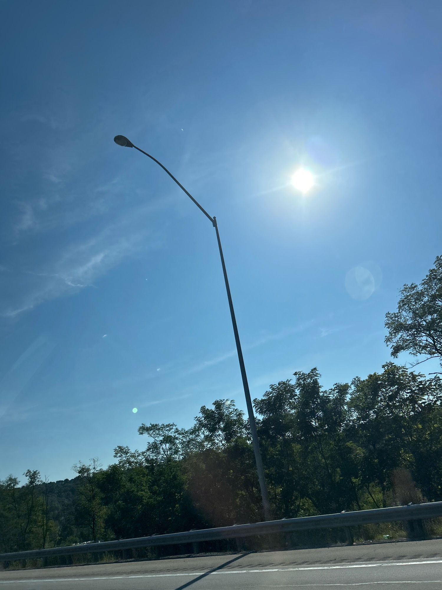 This is a photograph taken from a vehicle on a sunny day, looking towards a bright blue sky with some wispy clouds. A tall, dark street lamp with a curved arm extends from the bottom left of the frame upwards and to the right, its light fixture visible. The sun is shining brightly in the upper right quadrant, creating lens flare and washing out some of the sky. In the background, a dense line of green trees forms a natural barrier, with a subtle hint of hills or uneven terrain beyond them. A metal guardrail runs horizontally across the lower portion of the image, separating the road from the grassy shoulder and trees. The road itself is visible in the foreground, with white lane markings. The scene appears to be along a highway or major road, as suggested by the guardrail and the perspective from a moving vehicle. Based on the presence of this type of infrastructure and the natural landscape, the location is likely within a suburban or semi-rural area of Collier Township, United States of America. There are no people visible in the image.